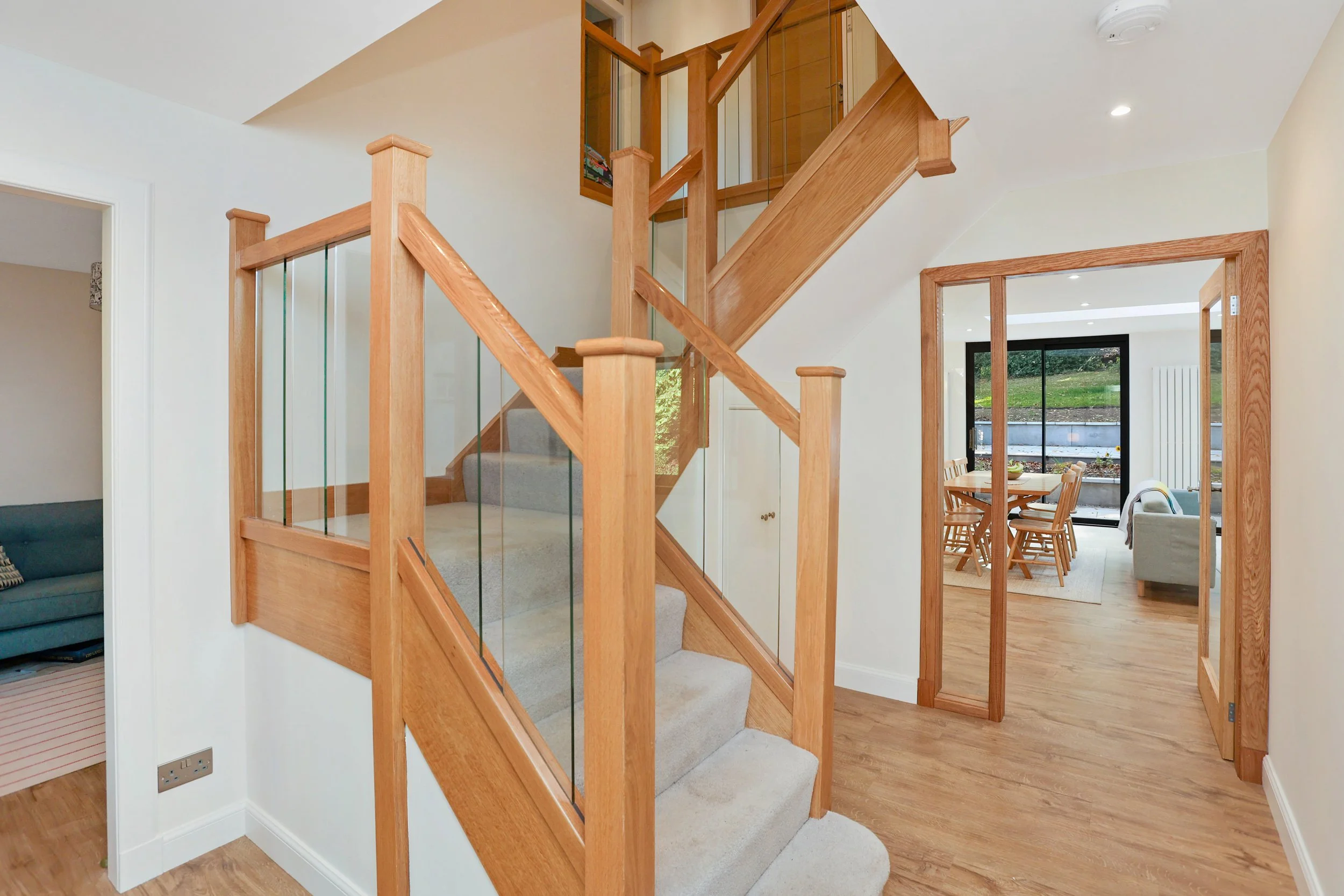 Interior view of a home staircase with wooden railings, glass panels, and carpeted steps, leading to an upper floor with a visible open corridor.