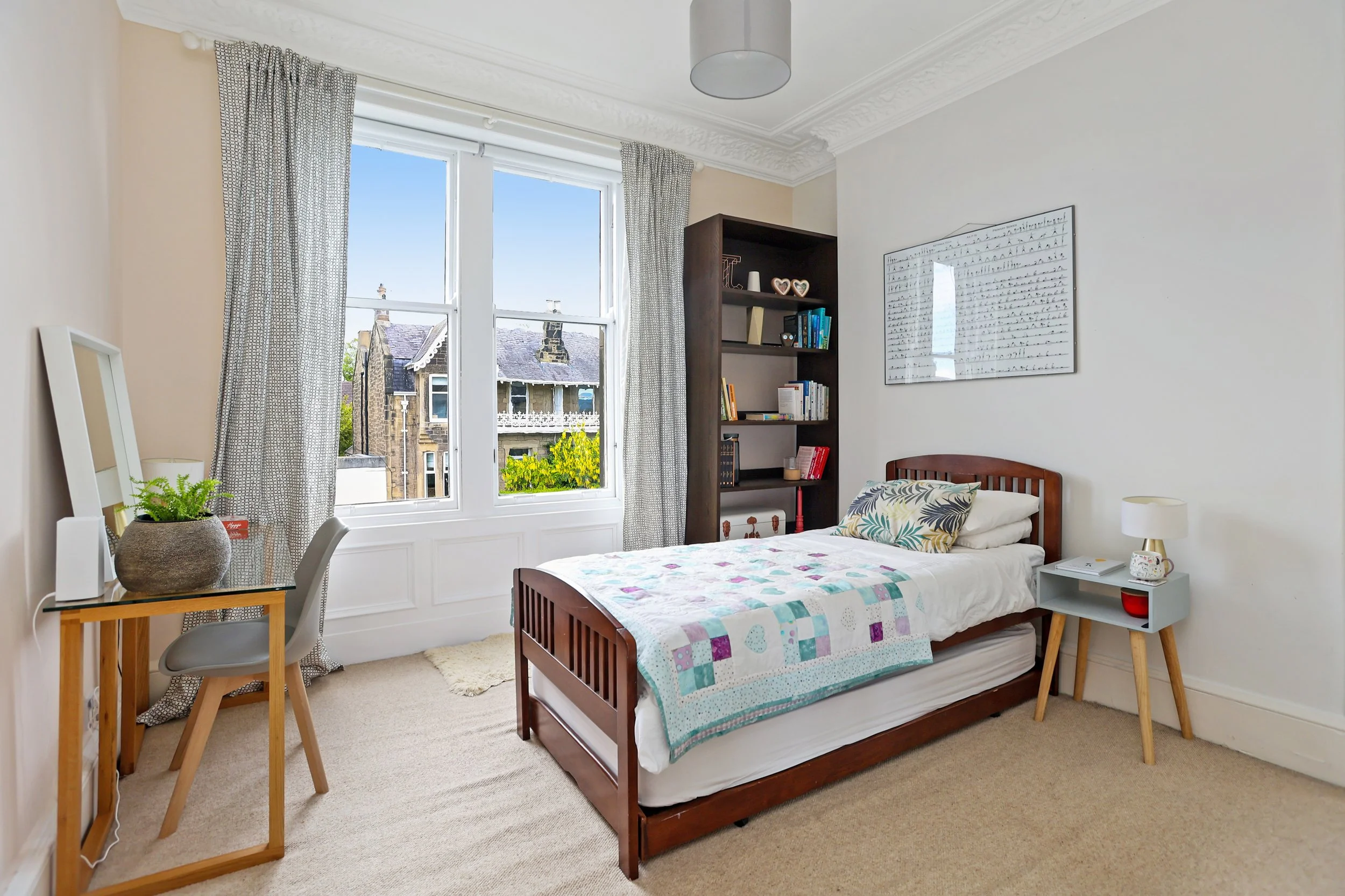 A bedroom with a single bed, a side table with a mug and a book, a bookshelf with books and decorative items, a large window with curtains showing rooftops and trees outside, a desk with a chair, and wall art with handwritten text.