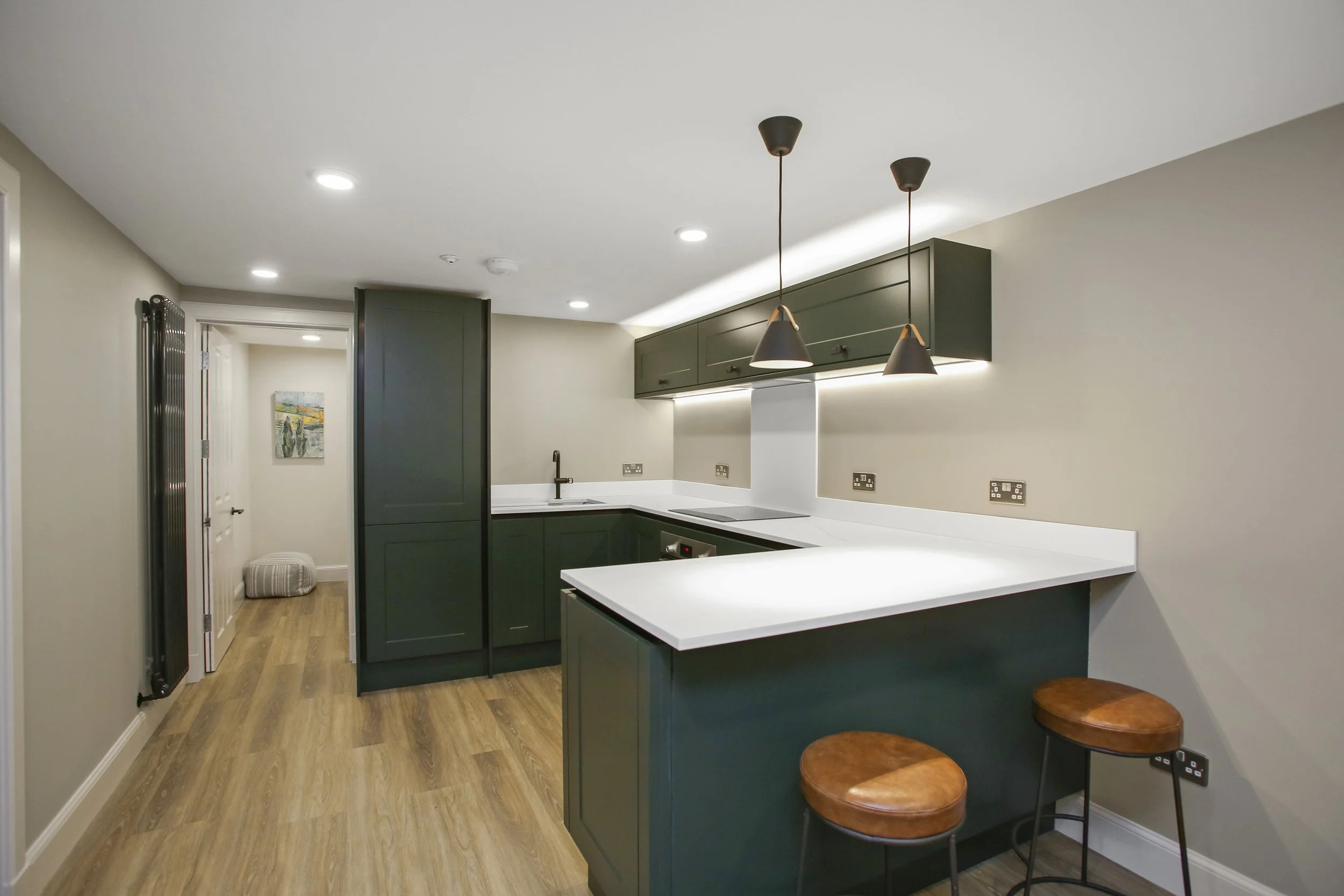 Modern kitchen with dark green cabinets, white countertops, pendant lighting, and a small breakfast bar with two wooden stools. The floor is wood, and there is a doorway to another room with a colorful painting on the wall.
