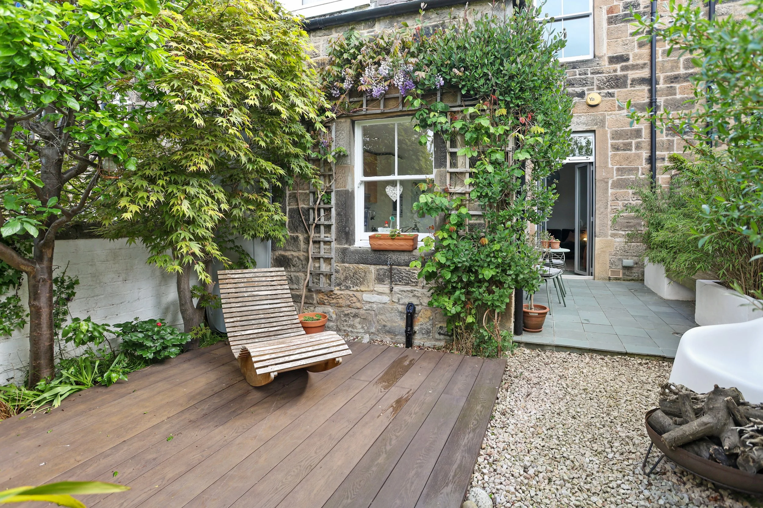 A cozy backyard patio with a wooden deck, green trees, potted plants, and a brick house with windows and an open door.