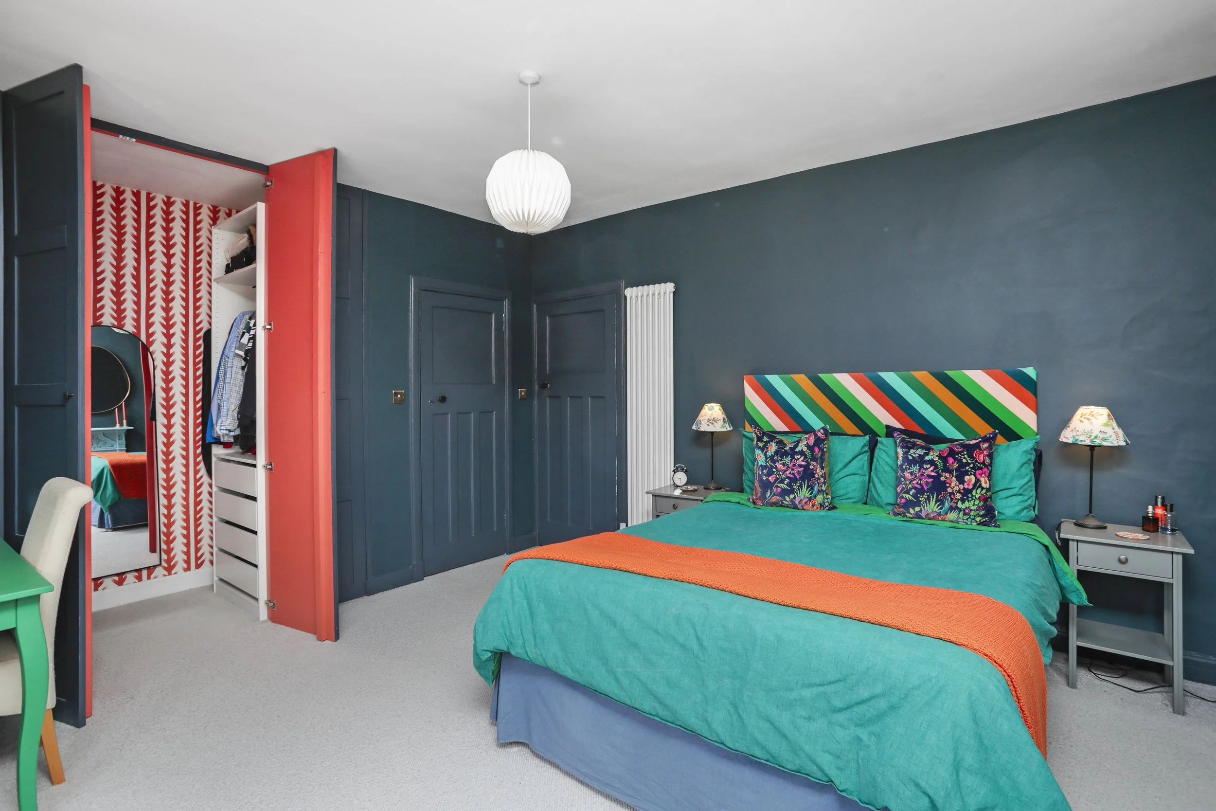 A colorful bedroom with teal walls and bedding, multicolored pillows, and bedside lamps. There is a white closet with red patterned sliding doors partially open, revealing hanging clothes inside. The room has a white ceiling with a hanging white pend