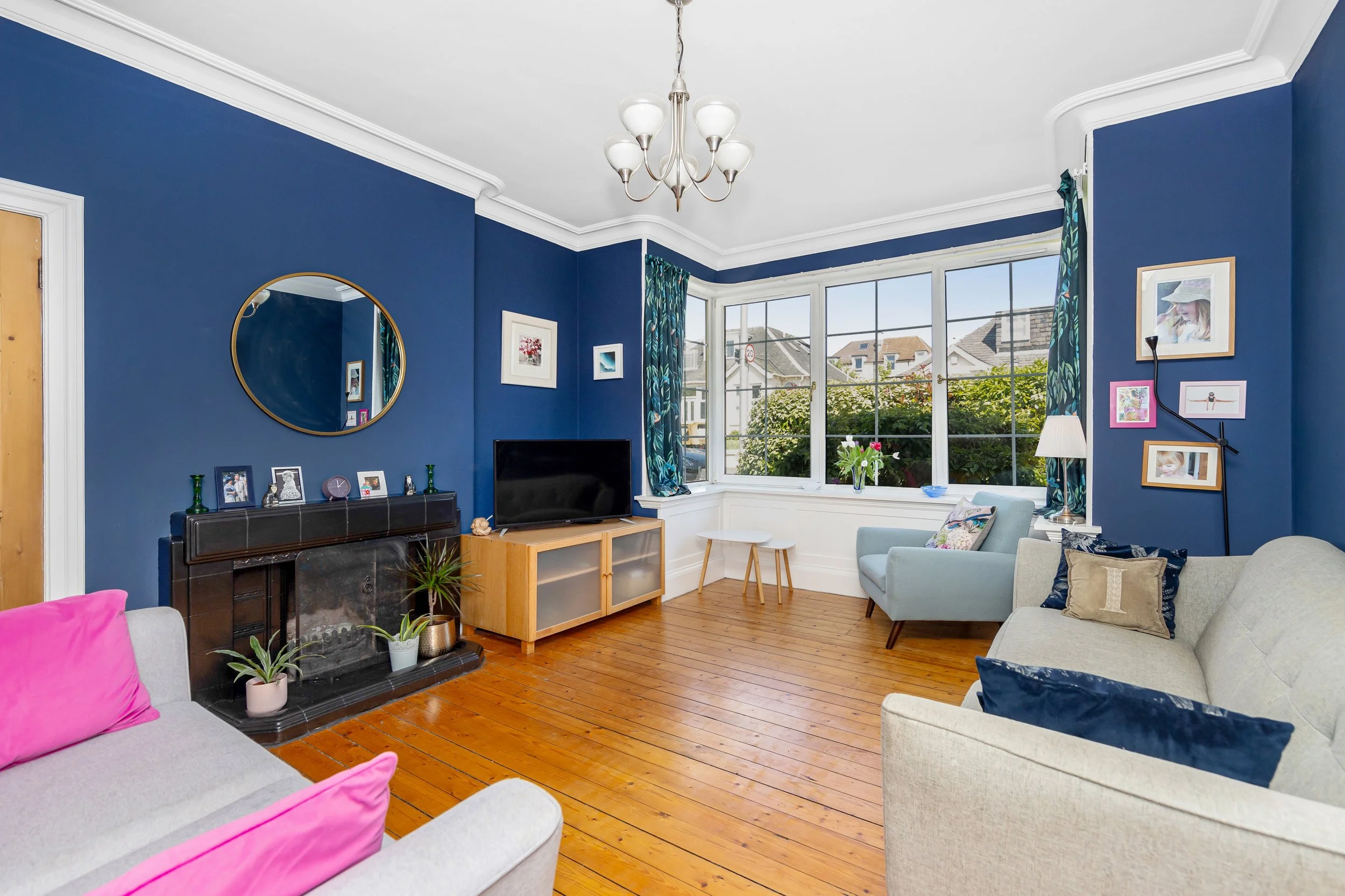 Living room with blue walls, wooden floor, a fireplace with a mirror above it, a television, a sofa with pink and blue pillows, a light green armchair, a window with curtains, framed pictures on the walls, and a chandelier.