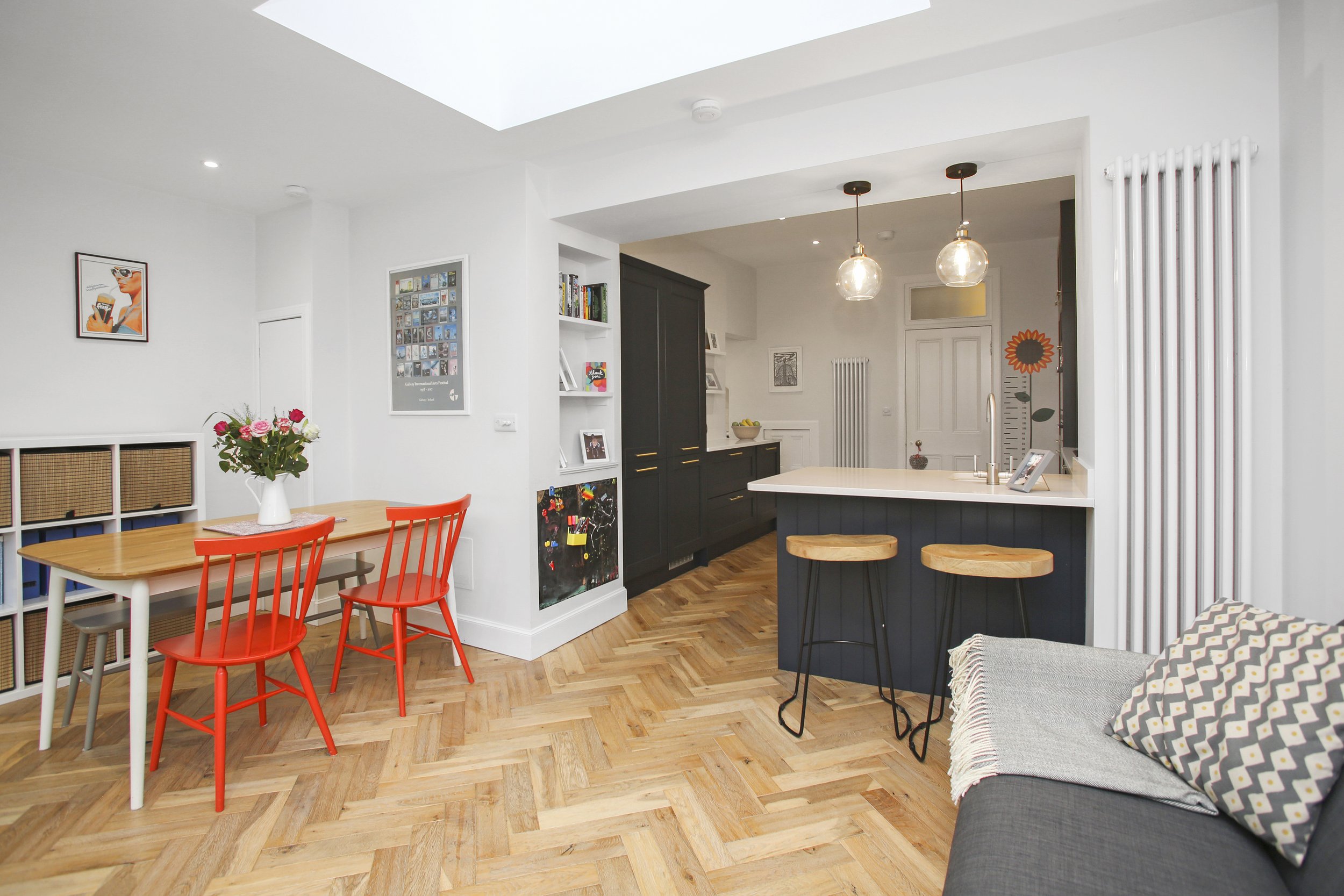 Open-concept living and dining area with a kitchen featuring black cabinets and a white island, a wooden dining table with red chairs, a gray sofa with patterned pillows, and wooden floors. Decor includes wall art, a plant, and pendant lights.