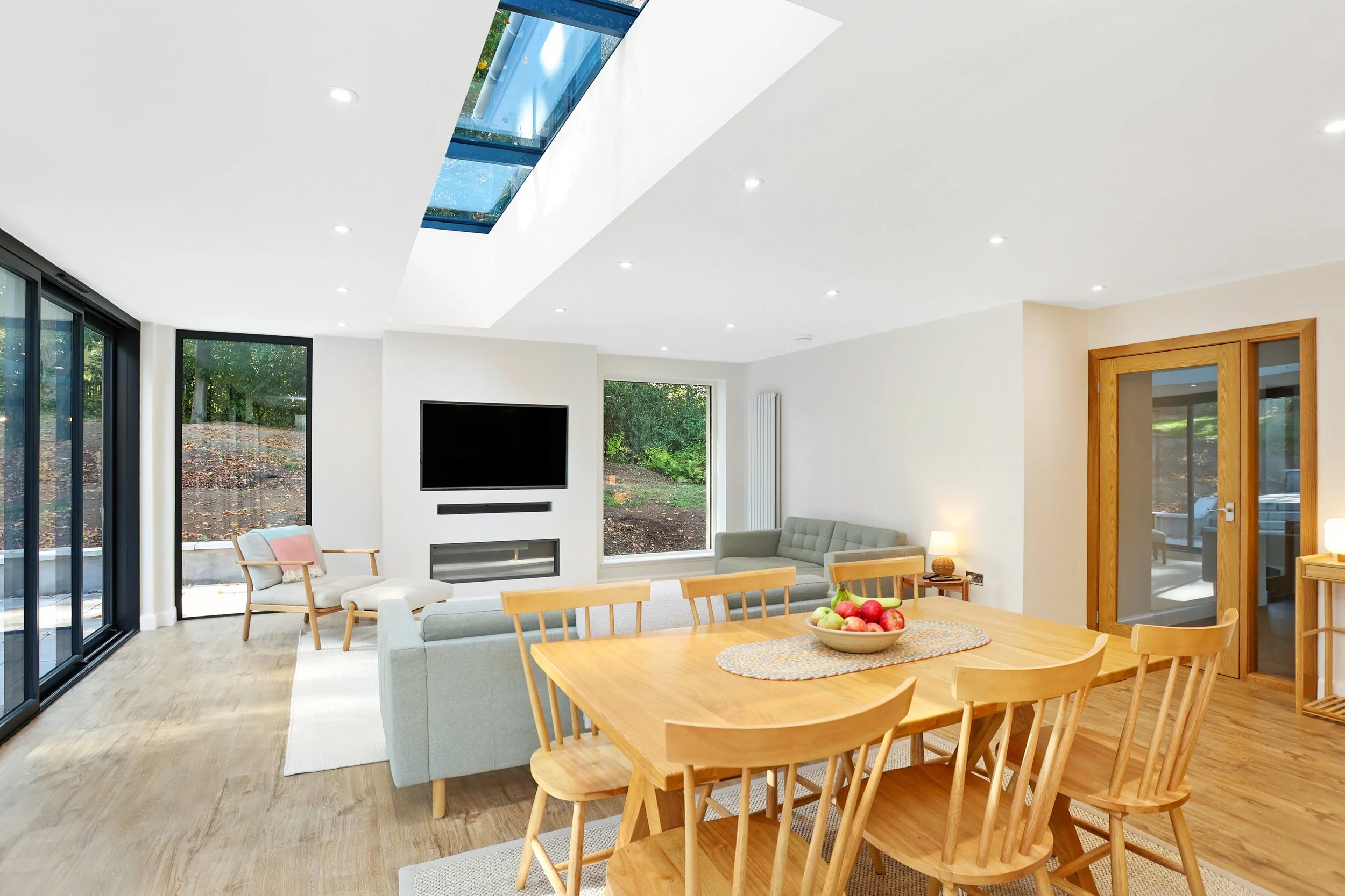 Open-concept living and dining room with large windows, skylight, and wooden furniture, including a dining table with a bowl of apples.