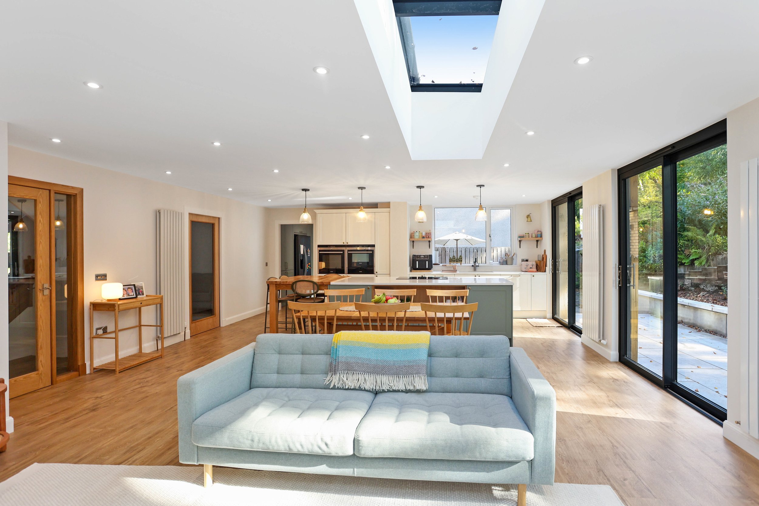 Open-concept kitchen and living area in a modern home with large sliding glass doors leading to an outdoor patio, vaulted ceiling with skylight, and hardwood floors. The kitchen features a white island and hanging pendant lights, while the living roo