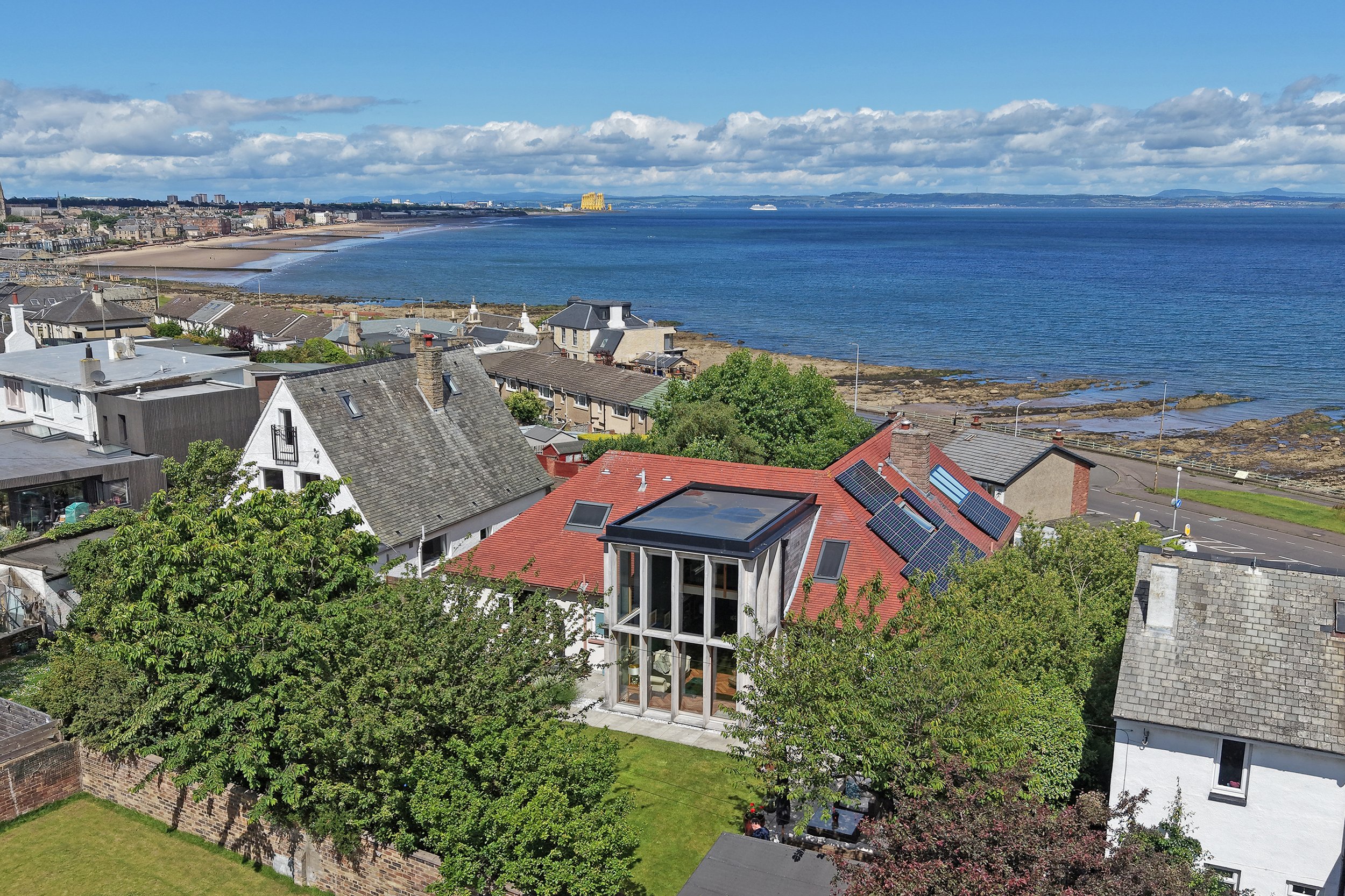 View from a high vantage point showing a coastal town with houses, trees, and rooftops in the foreground, a beach and ocean in the middle distance, and a cloudy sky with blue patches in the background.