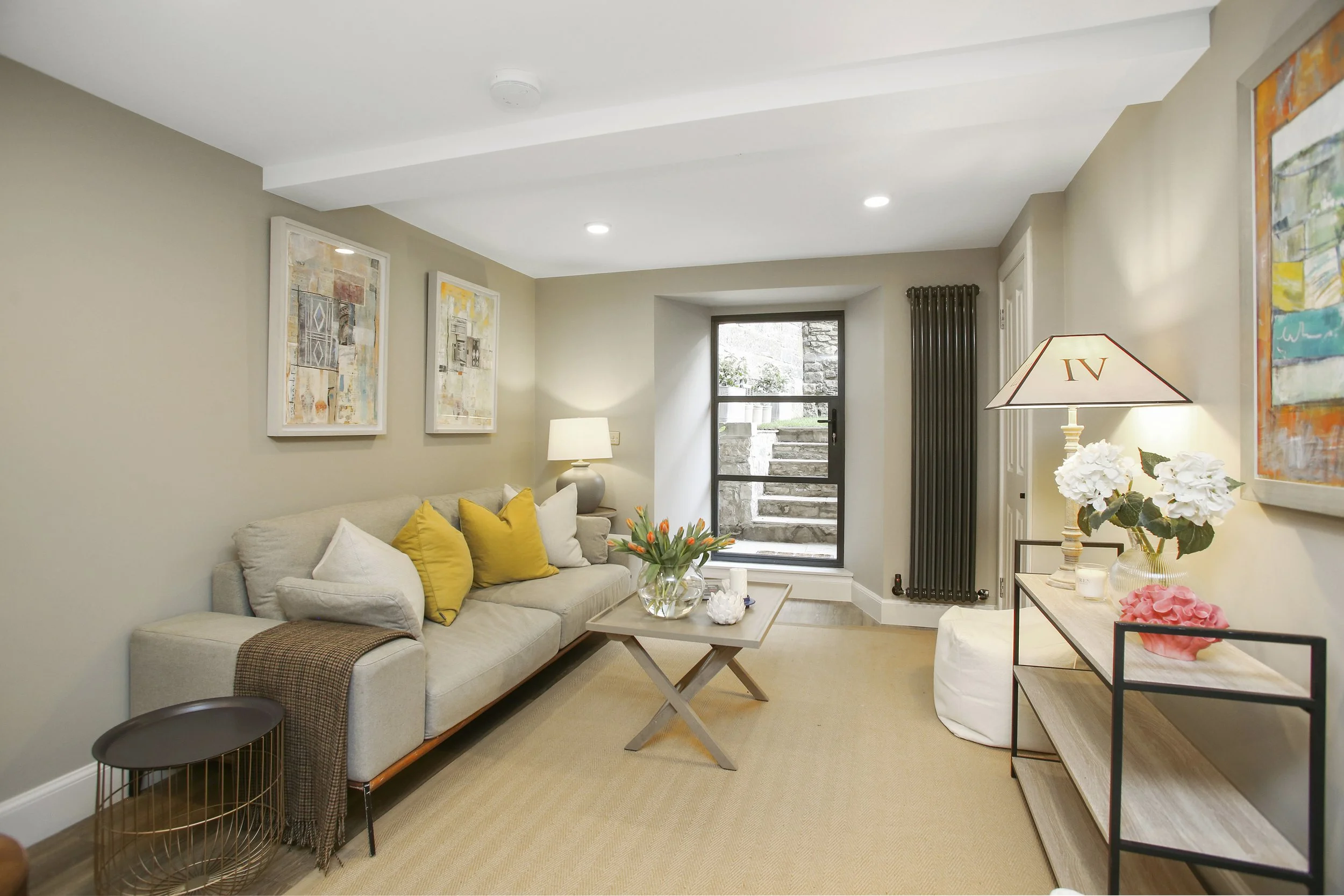 Living room with beige walls, cream sofa with yellow and white pillows, wooden coffee table with flowers, abstract artwork on walls, window with stone wall outside, radiator, white side table with flowers and lamp.