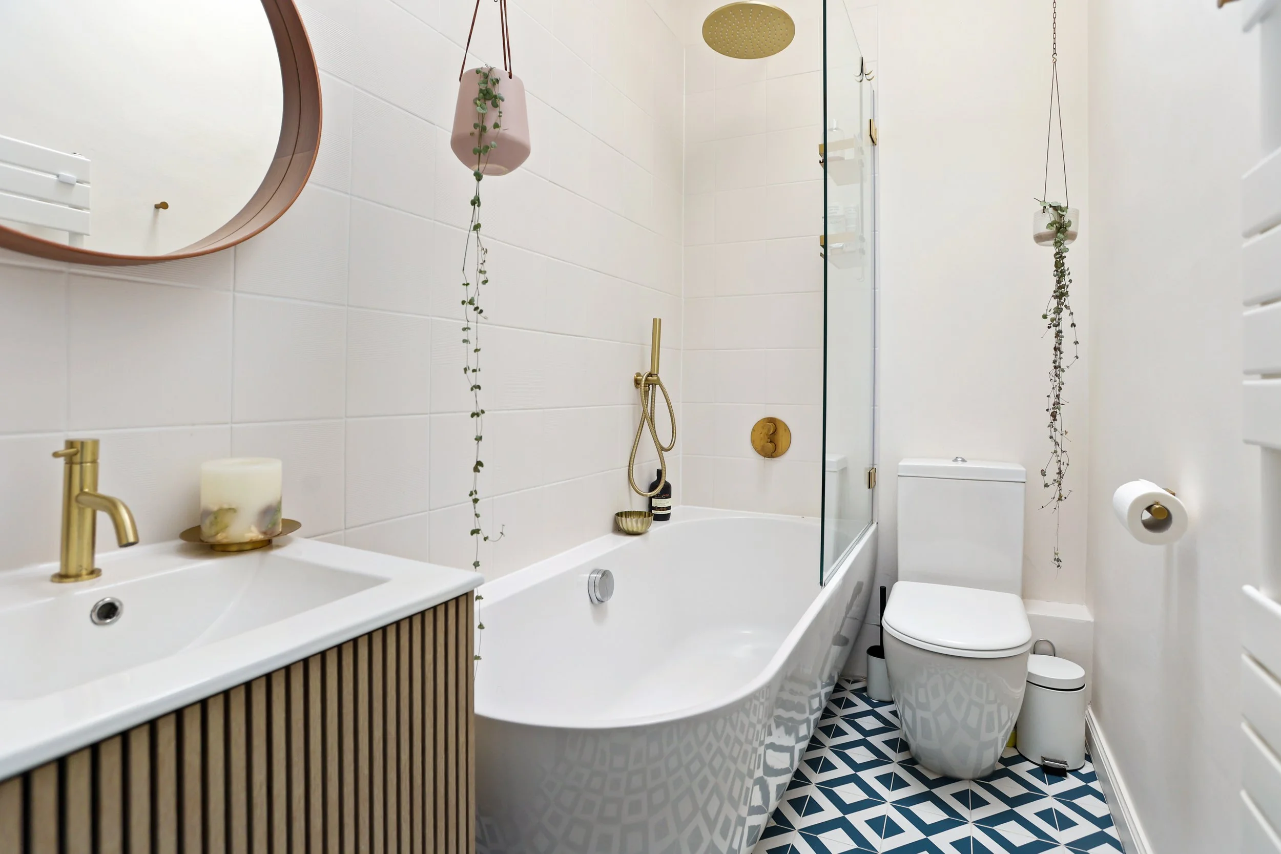 A modern bathroom with a white bathtub, gold hardware, hanging plants, patterned floor tiles, a mirror, and a toilet.