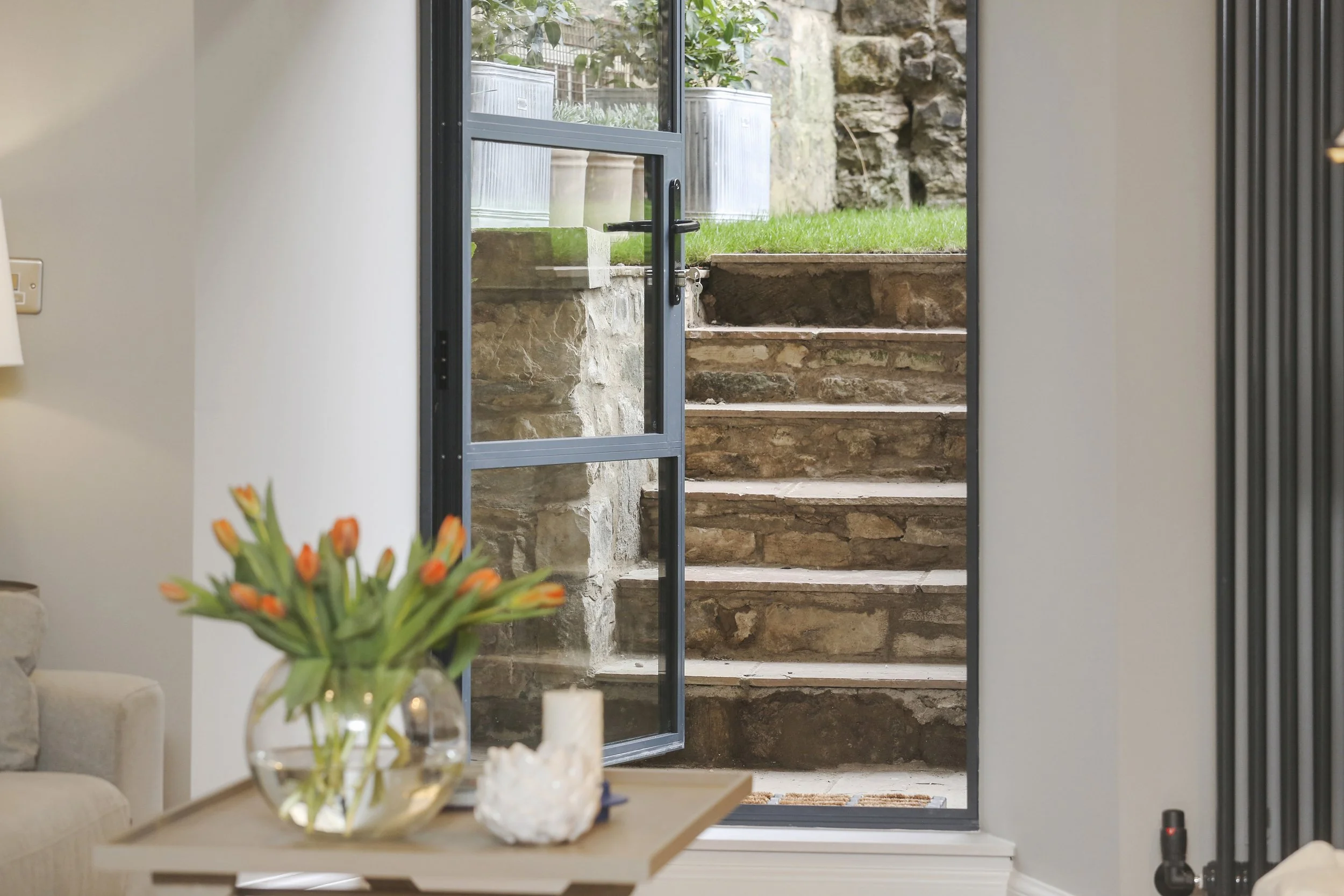 Interior view of a living room with a glass door leading outside to a garden with stone steps, grass, and large outdoor planters.