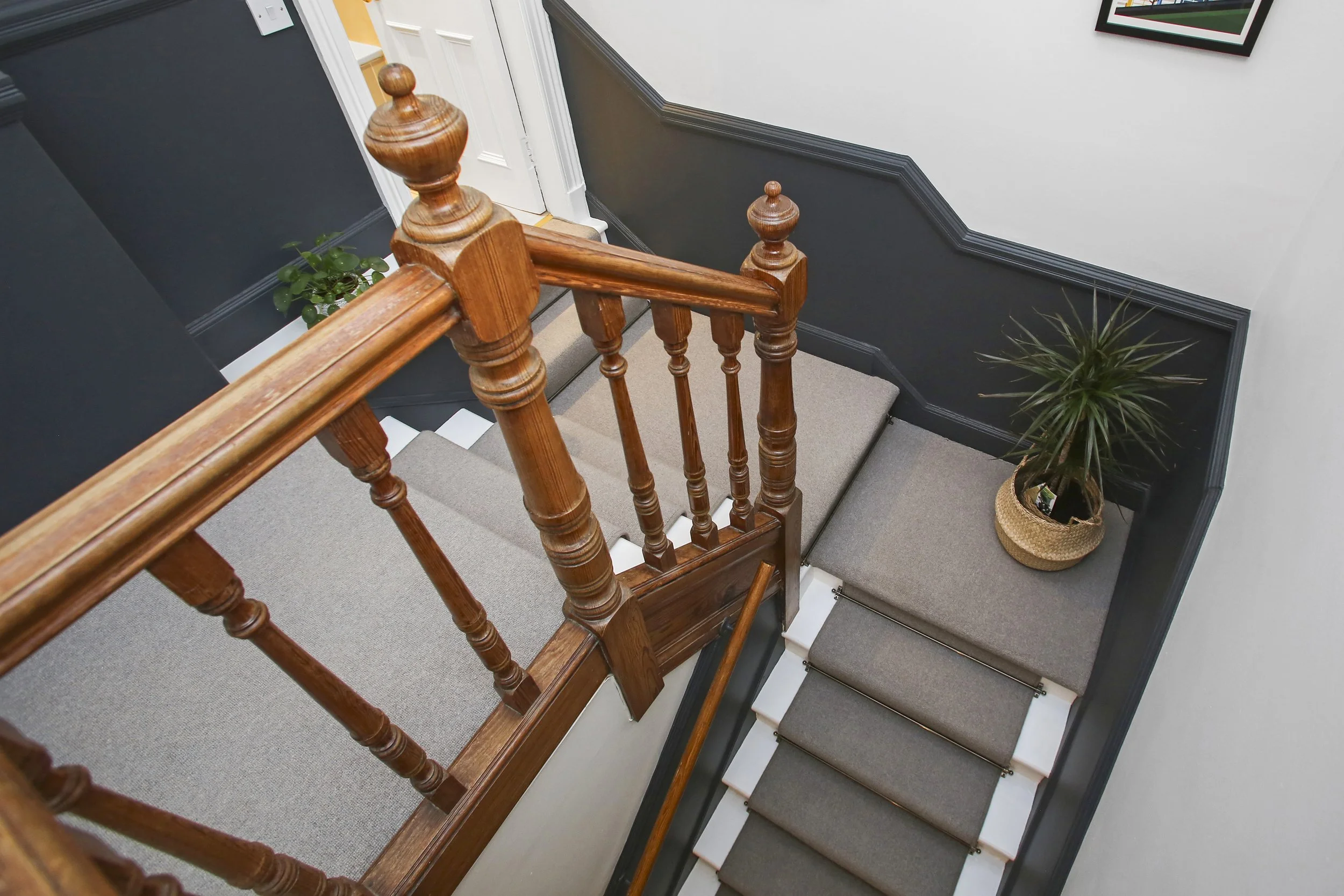 Inner view of a staircase with a wooden handrail and spindles, gray wall with black trim, and potted plants. There is a door at the top of the stairs.