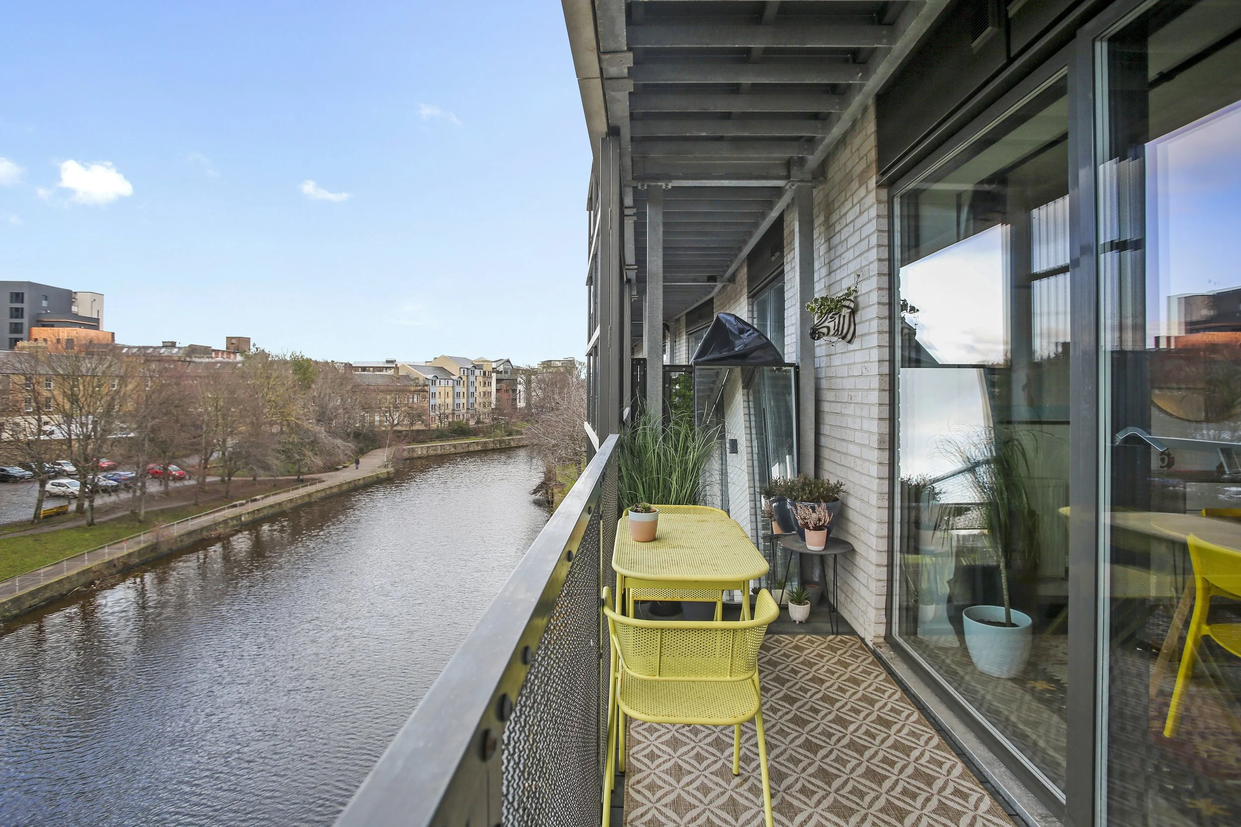Balcony with yellow chairs and potted plants overlooking a waterway with trees and buildings