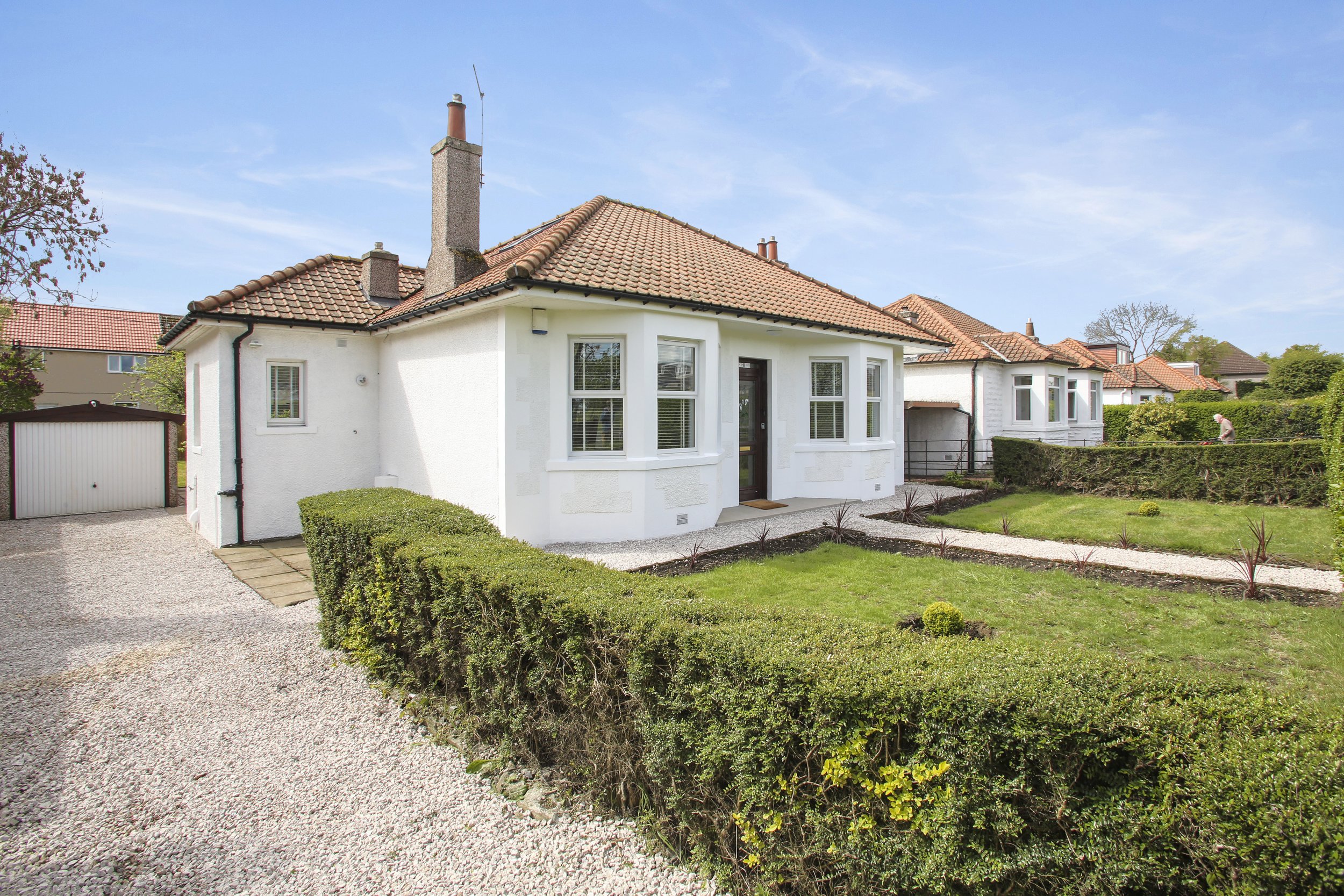 A white house with red tile roof, surrounded by a gravel driveway and neatly trimmed bushes and lawn, on a sunny day with blue sky.