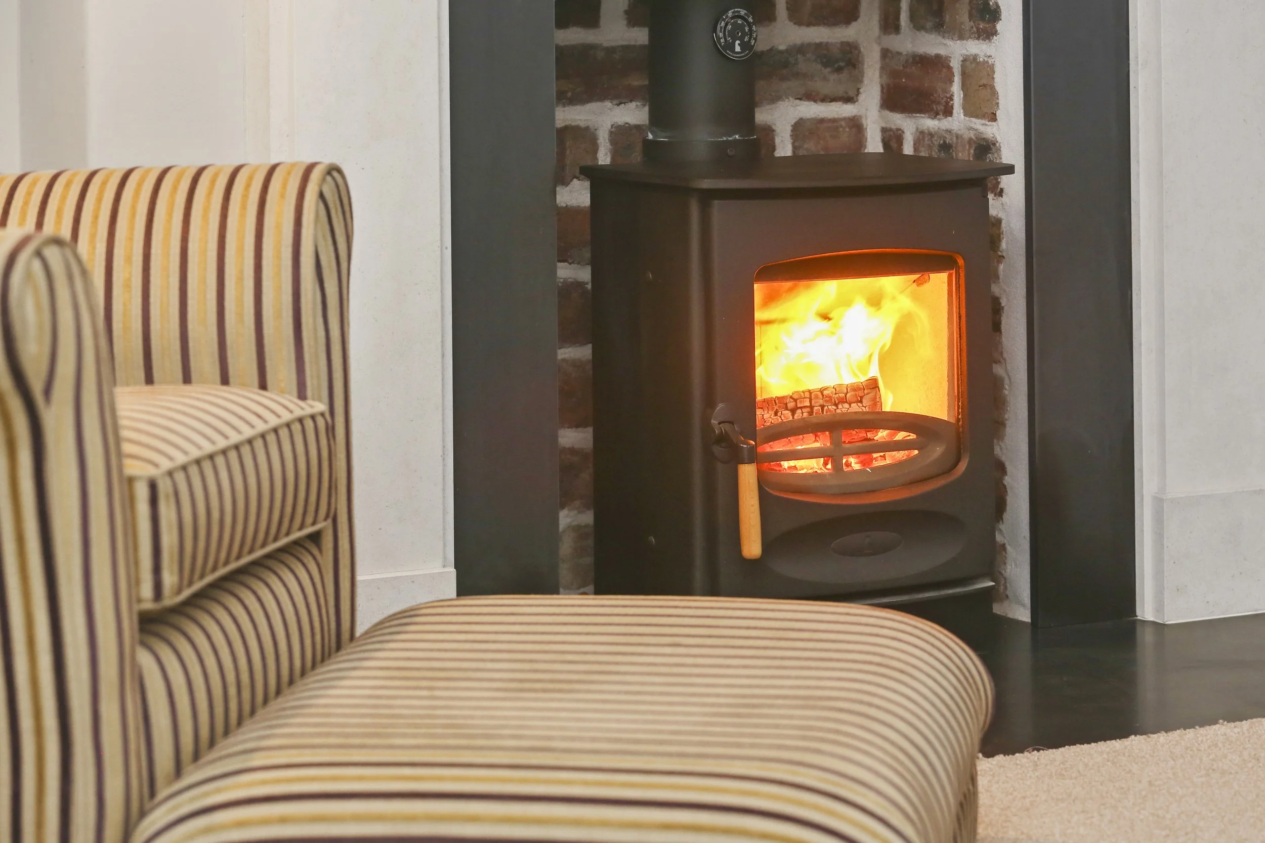 A living room corner featuring a wood-burning stove with an active fire, brick wall background, striped upholstered armchair, and a beige carpeted floor.