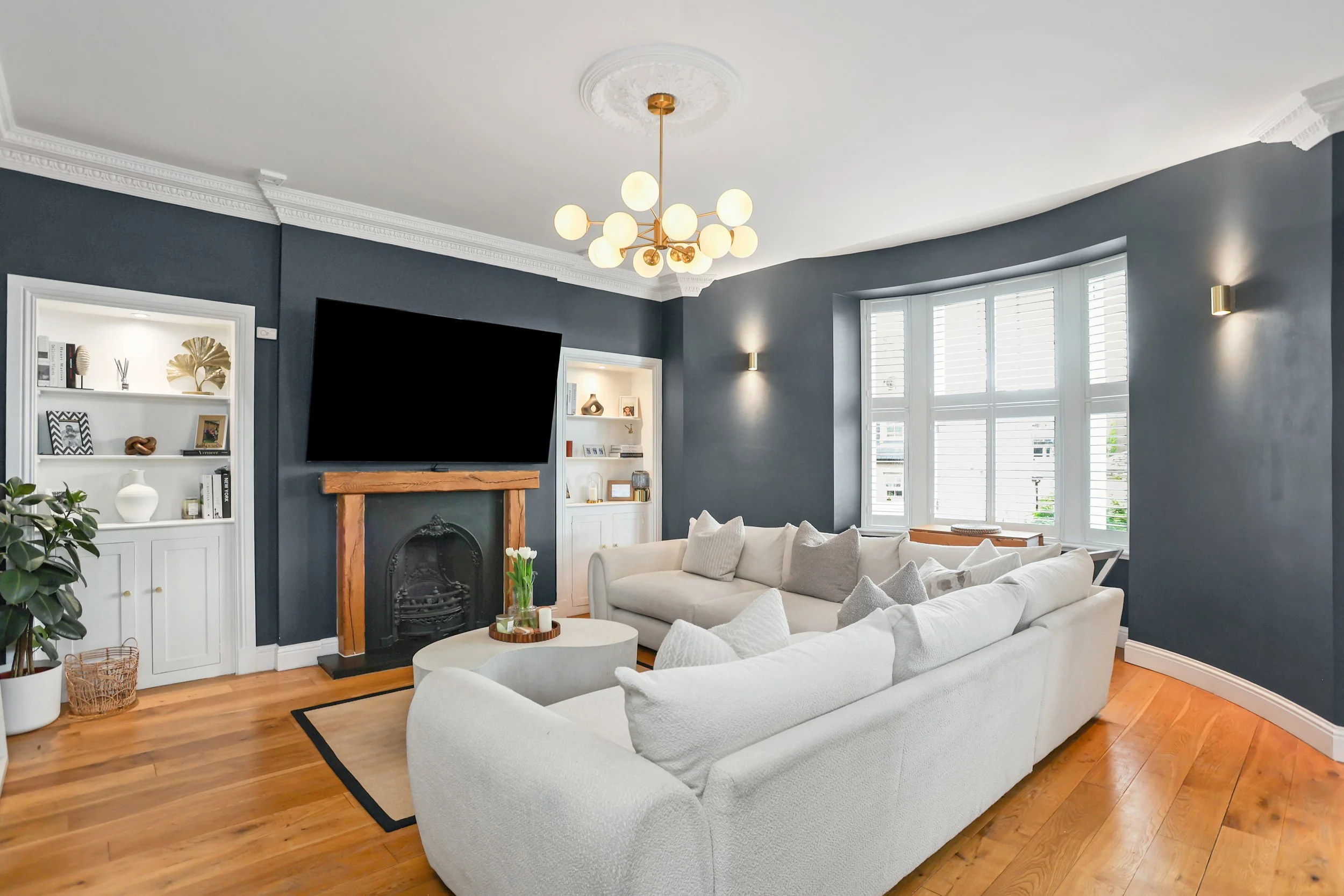 Living room with dark gray walls, white crown molding, large window with white shutters, white sofa, round coffee table with flowers, wall-mounted TV above a fireplace with a wooden mantel, built-in white shelves with decorative items, wood flooring,