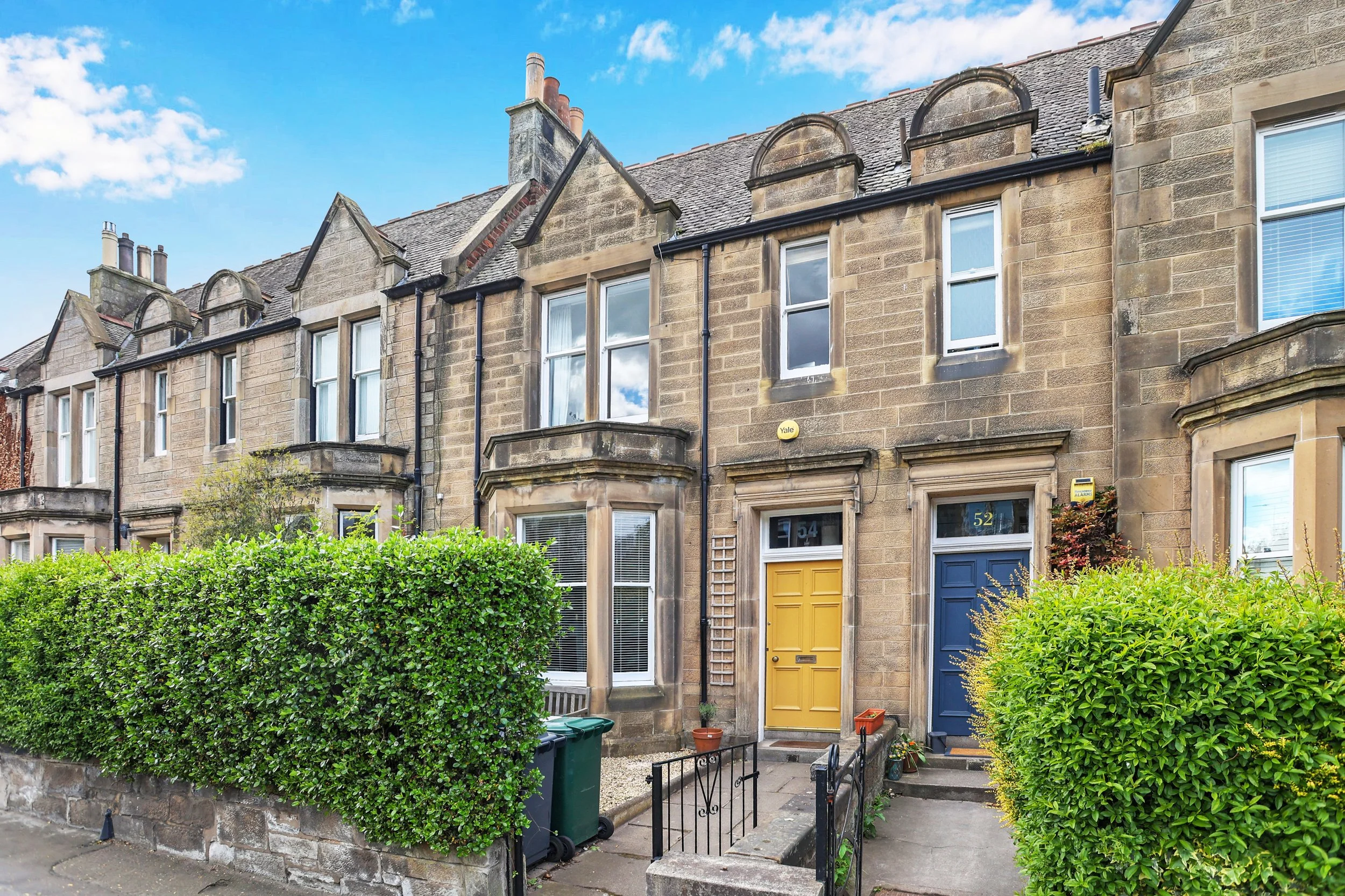 Row of traditional sandstone townhouses with colorful front doors, front gardens, and satellite dishes on the roofs, under a blue sky with scattered clouds.