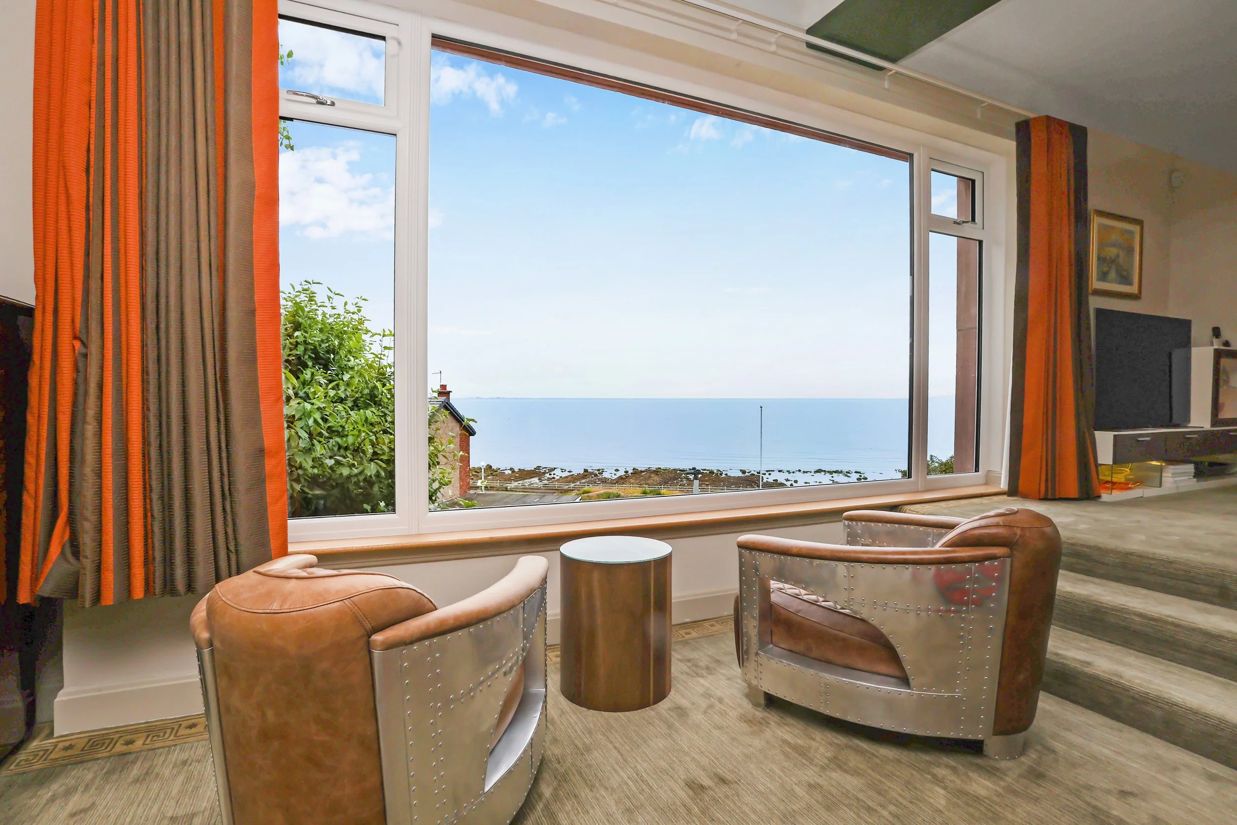 A living room with large window showing a view of the ocean, blue sky, and clouds, decorated with orange and beige curtains, chairs, a small round table, and a television.