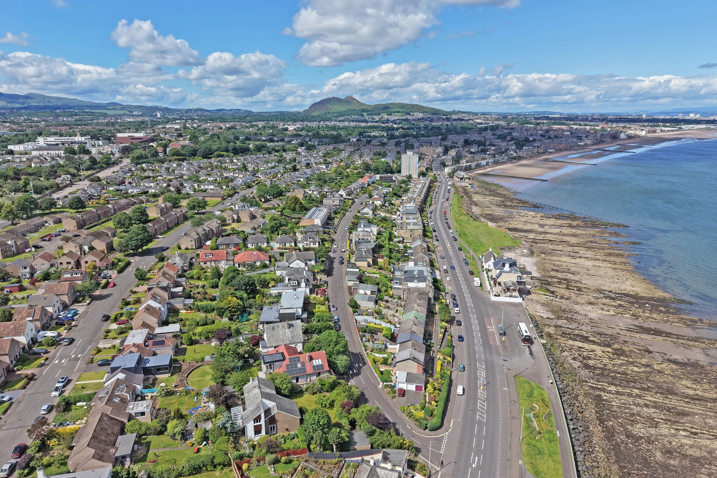 An aerial view of a coastal town with numerous houses, a road running parallel to the shoreline, and a shoreline with rocks and water. In the background, there are green hills and a partly cloudy sky.