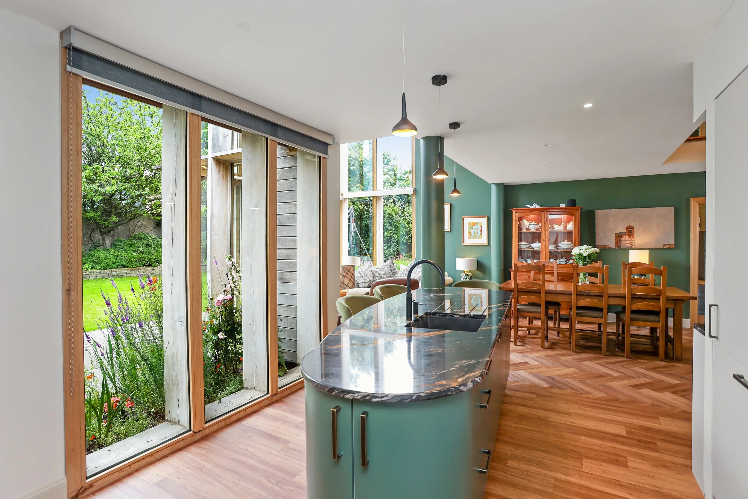 Modern open-concept kitchen with a view of a lush, green garden through large sliding glass doors. The kitchen features a black marble island with a built-in sink, green cabinetry, and wooden flooring. In the background, there is a wooden dining tabl