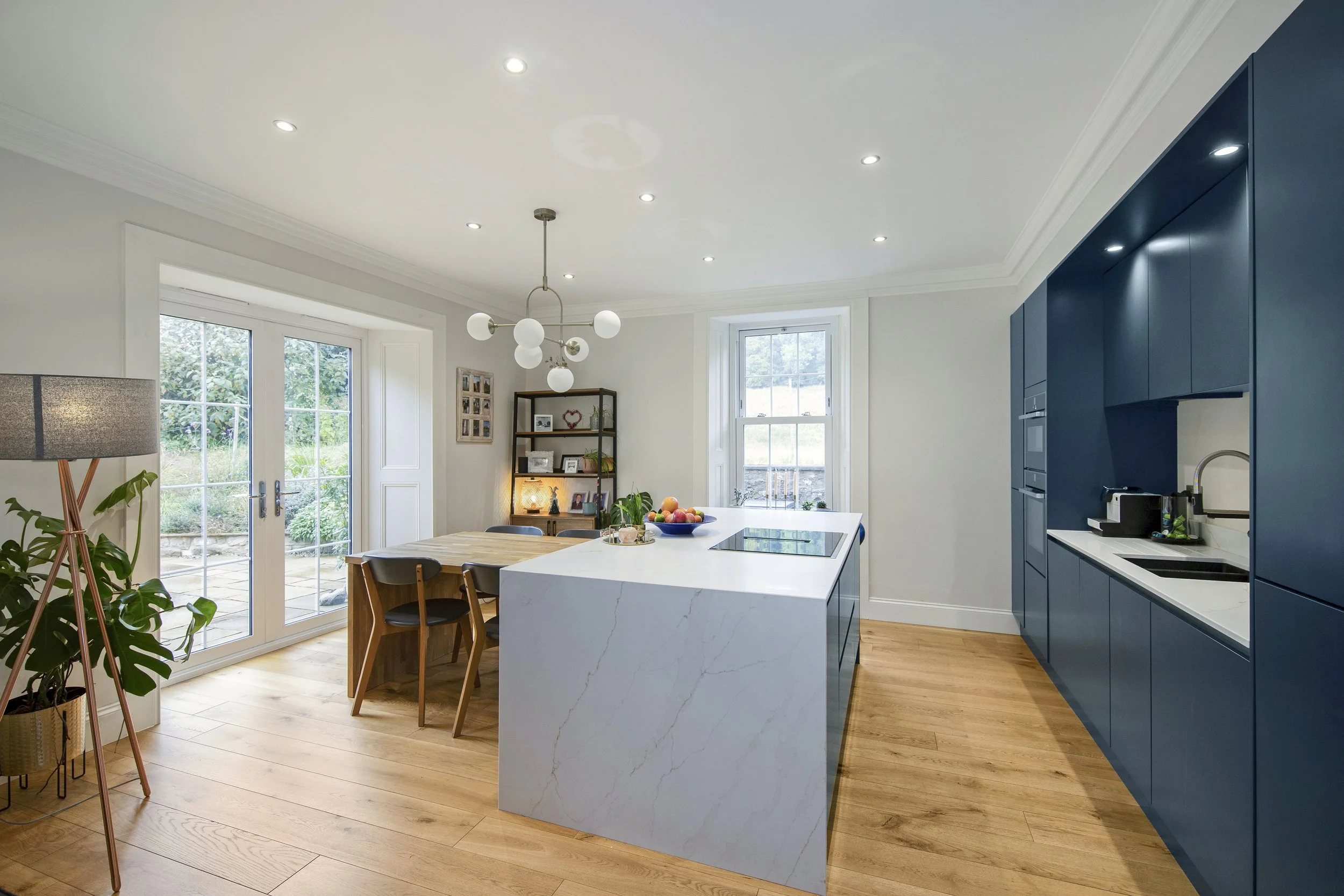 Modern kitchen with white countertops, blue cabinets, wooden flooring, and large windows showing greenery outside.