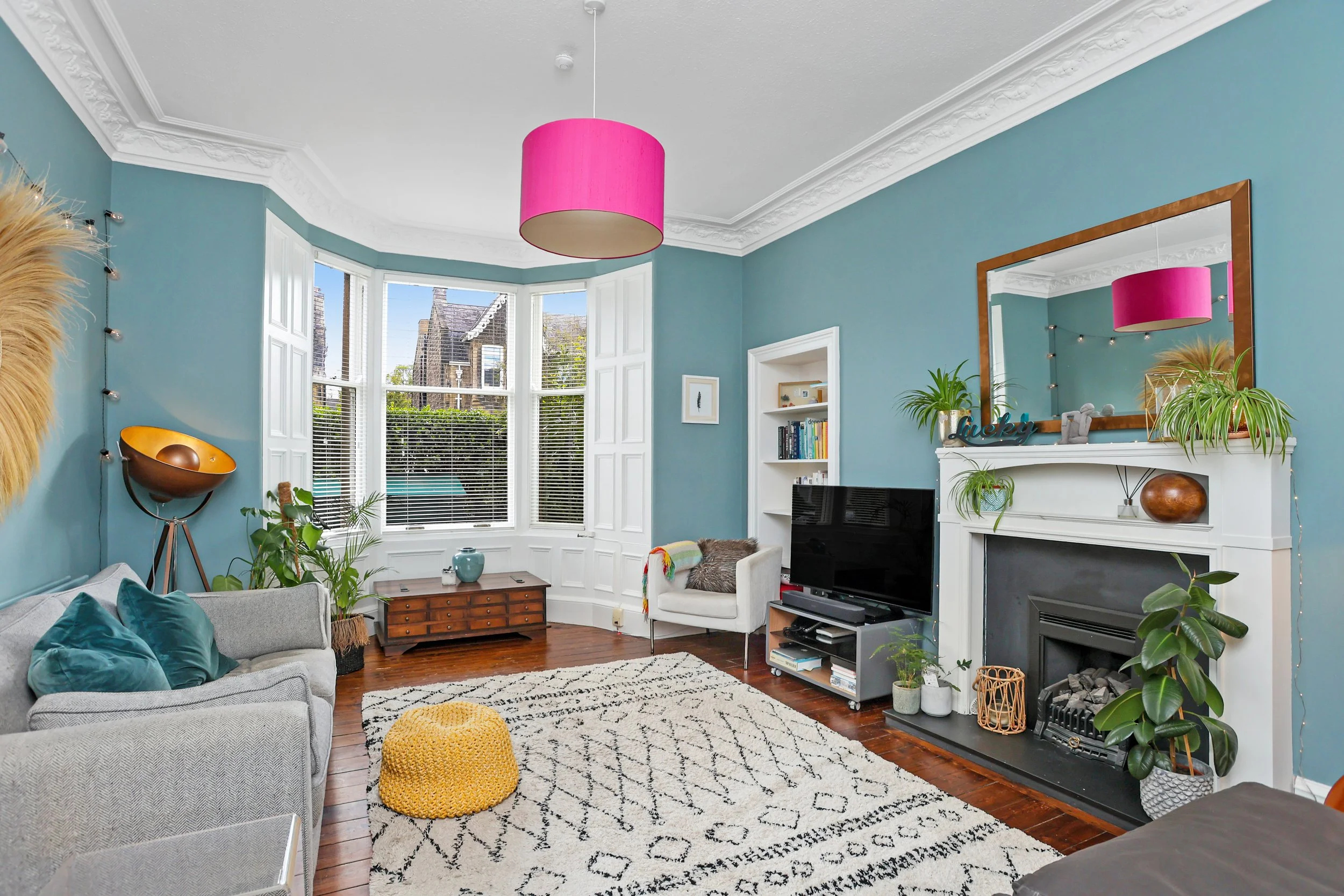 Living room with light blue walls, large bay window with white trim, and a white fireplace with a large mirror above. The room has a gray sofa with teal pillows, a white armchair, a small wooden coffee table, and various houseplants. A pink ceiling l