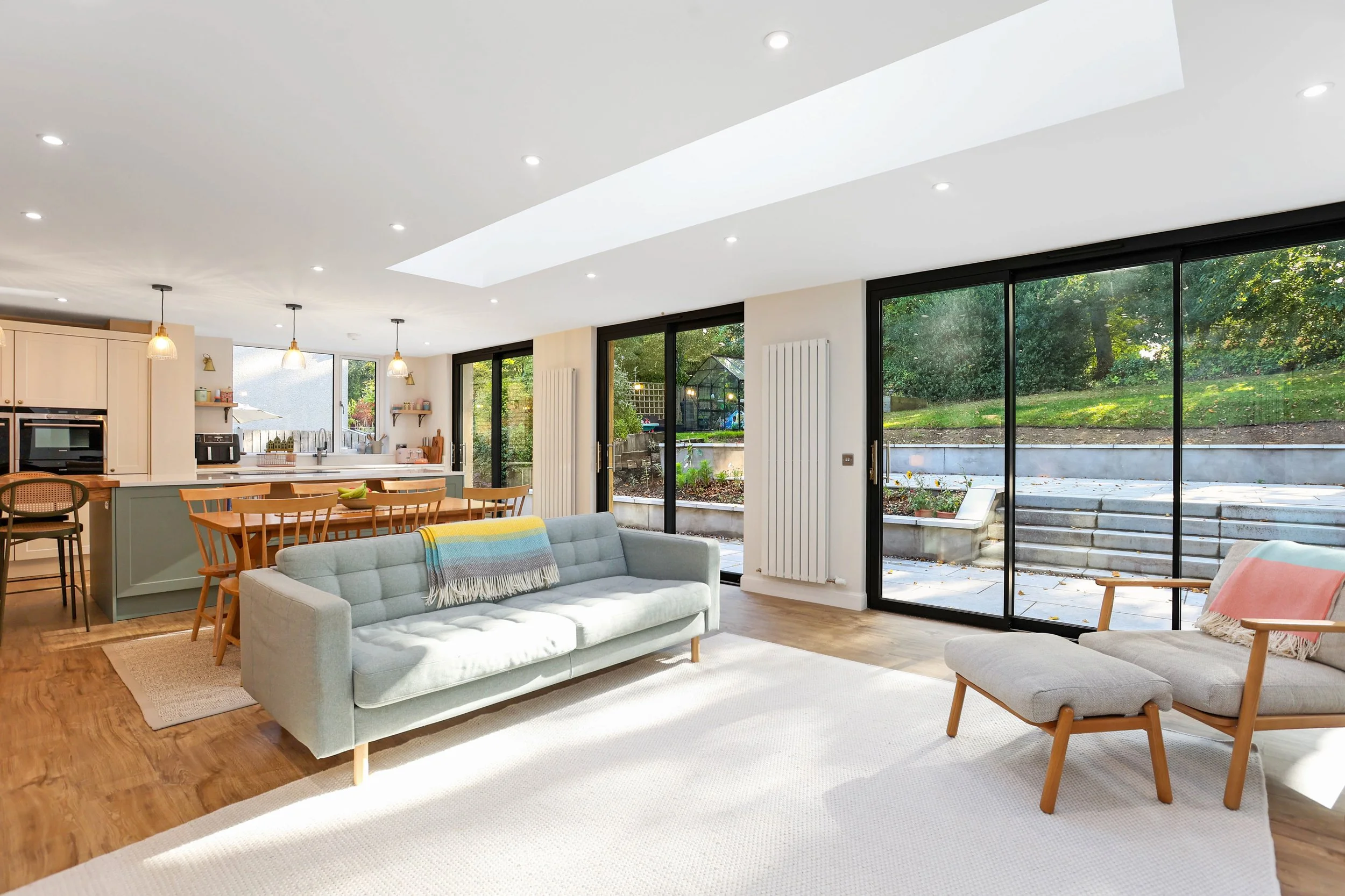 A modern open-plan living space with large floor-to-ceiling windows, a white couch with colorful throw, a light blue armchair, a wooden dining table with chairs, a kitchen area with pendant lights, and a view of a garden outside.