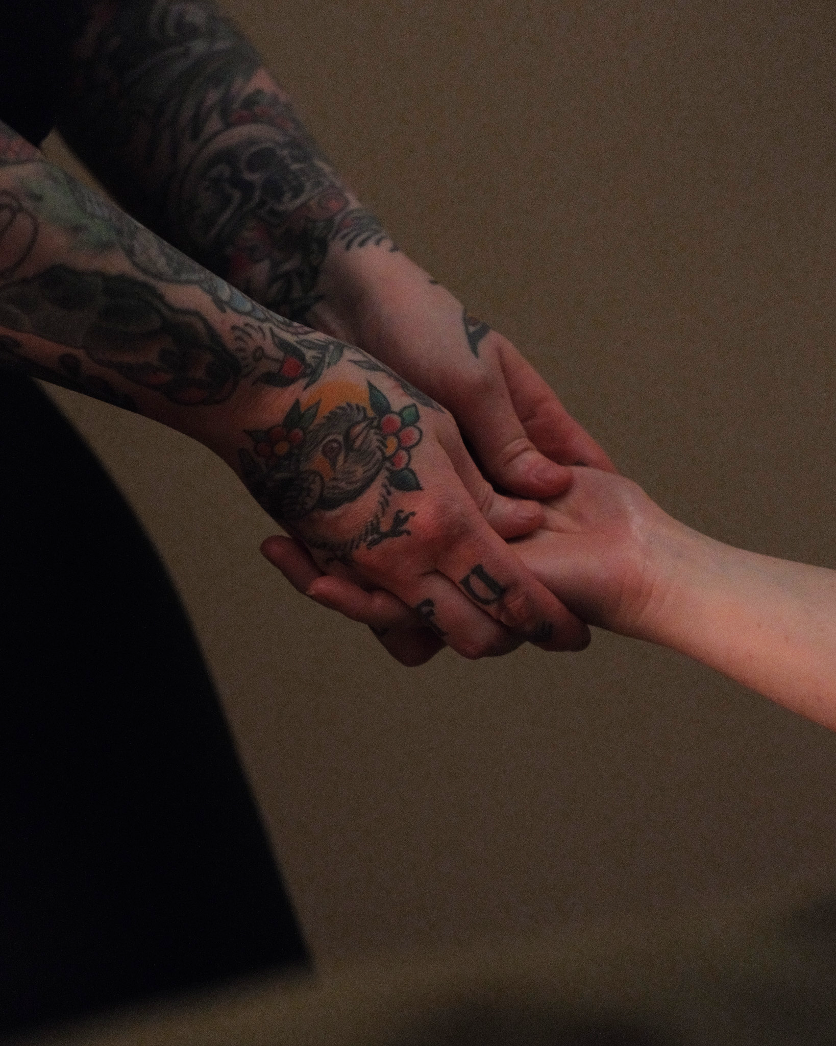 Two tattooed arms reaching to clasp hands, with a plain beige background.