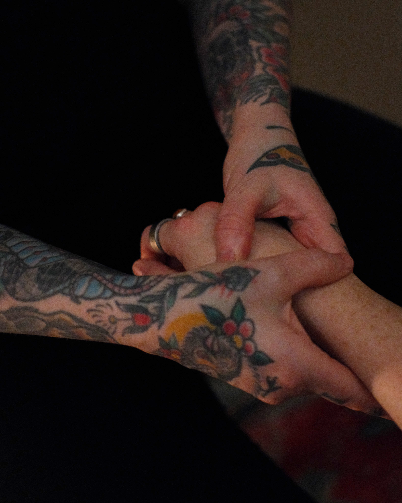 Close-up of two tattooed hands holding each other, with tattoos on arms and hands, and rings on fingers.