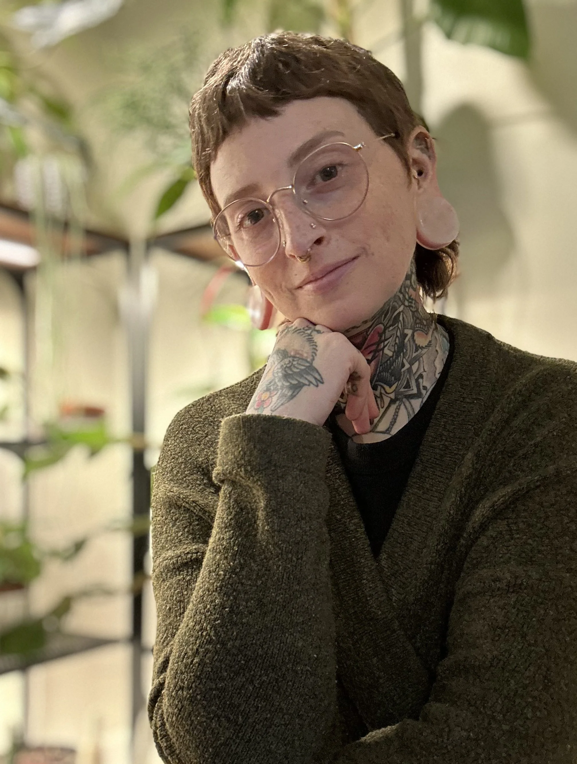 A person with short, dark hair, glasses, and facial tattoos resting their chin on their hand in an indoor setting with plants in the background.