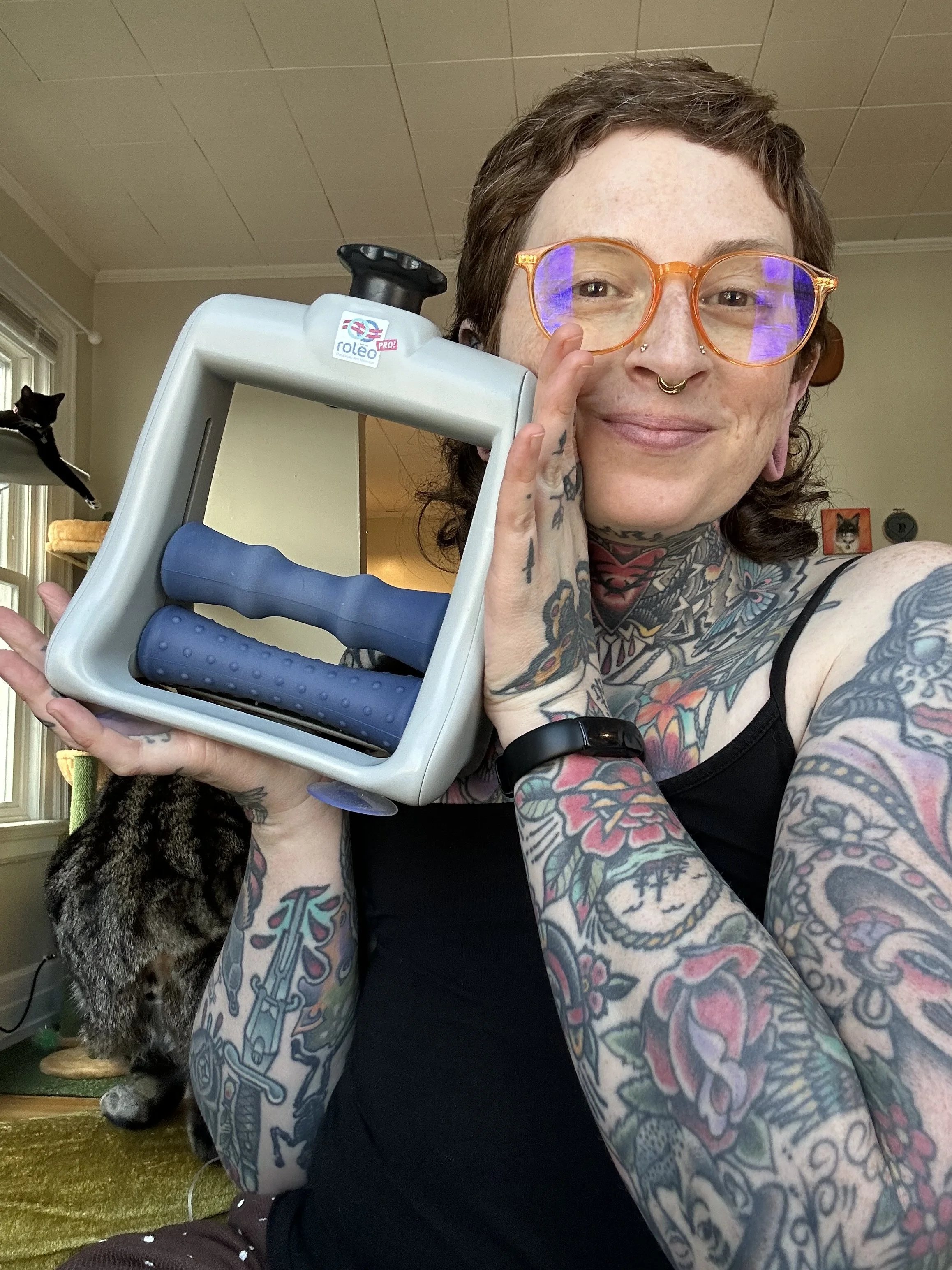 A person with tattoos wearing orange glasses, holding a recovery tool with blue handle grips, sitting indoors with a black cat in the background.