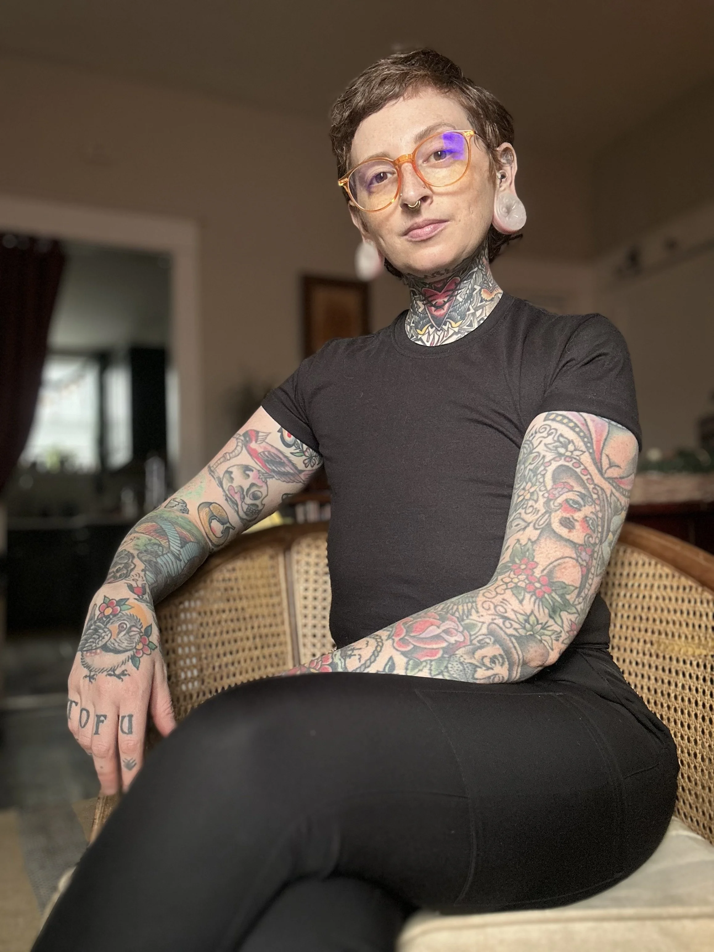 A person with short, dark hair, wearing orange glasses and tattoos on arms and neck, sitting on a wicker chair in a room with brown walls and a window in the background.