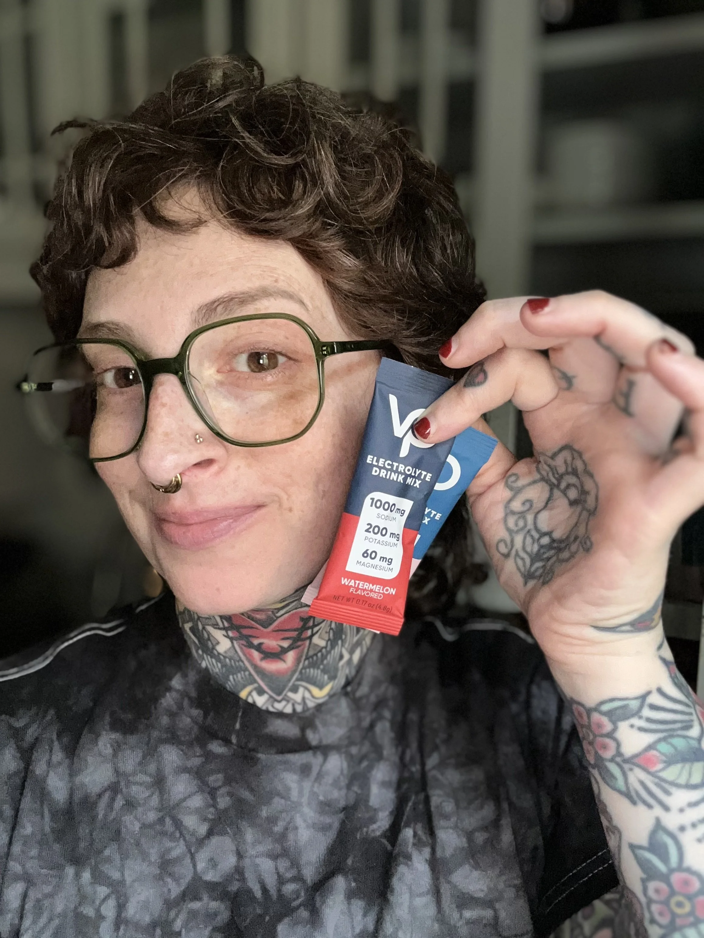 A person with curly brown hair, glasses, and tattoos on their arms is holding a blue packet of electrolyte drink mix labeled watermelon flavor, near their face. The person has a nose piercing and a tattoo on their chest. The background appears to be a kitchen or pantry with shelves.