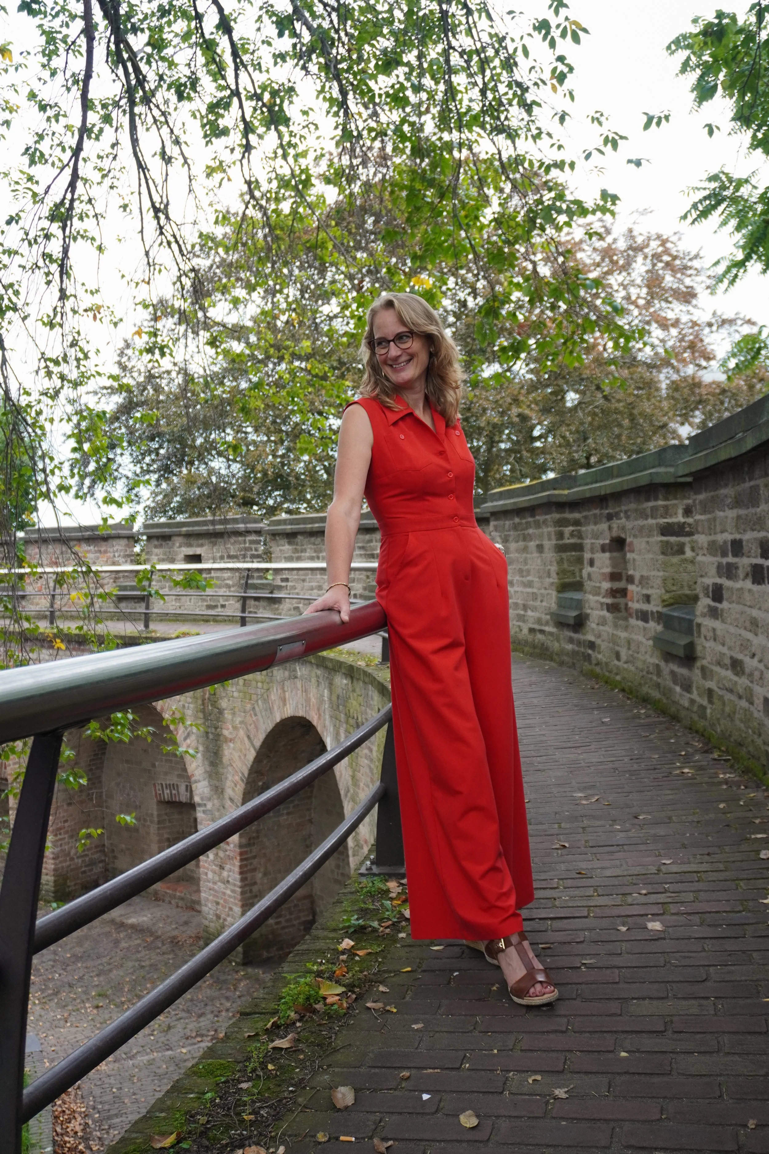 Een vrouw in een rode jumpsuit poseert op een brug met smeedwerk en stenen muur op de achtergrond, omringd door groene bladeren en bomen.
