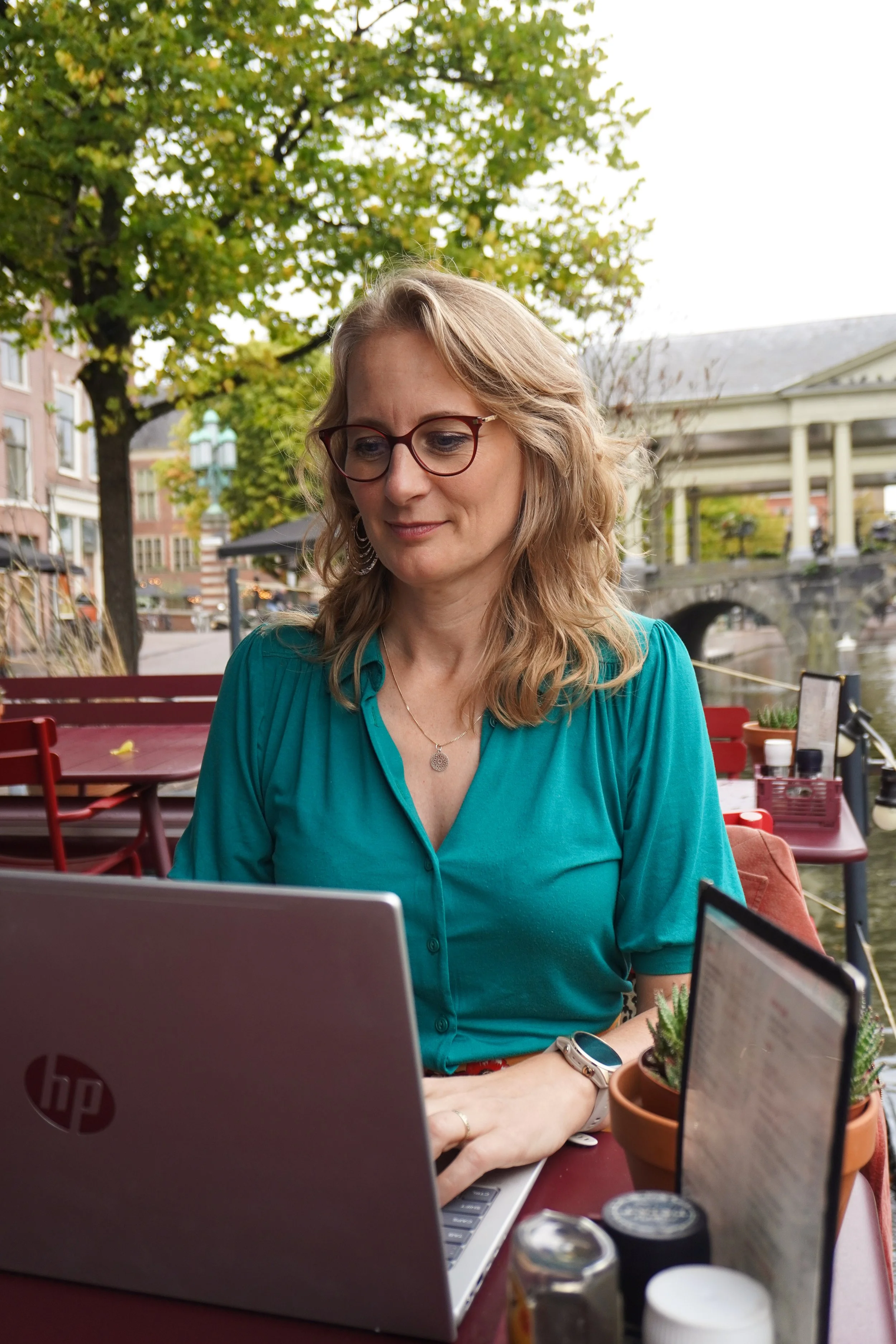 Vrouw met blonde haar en brilmontuur werkt op haar laptop op een terras met een rivier en brug op de achtergrond, omgeven door groene bomen, onder een overkapping.