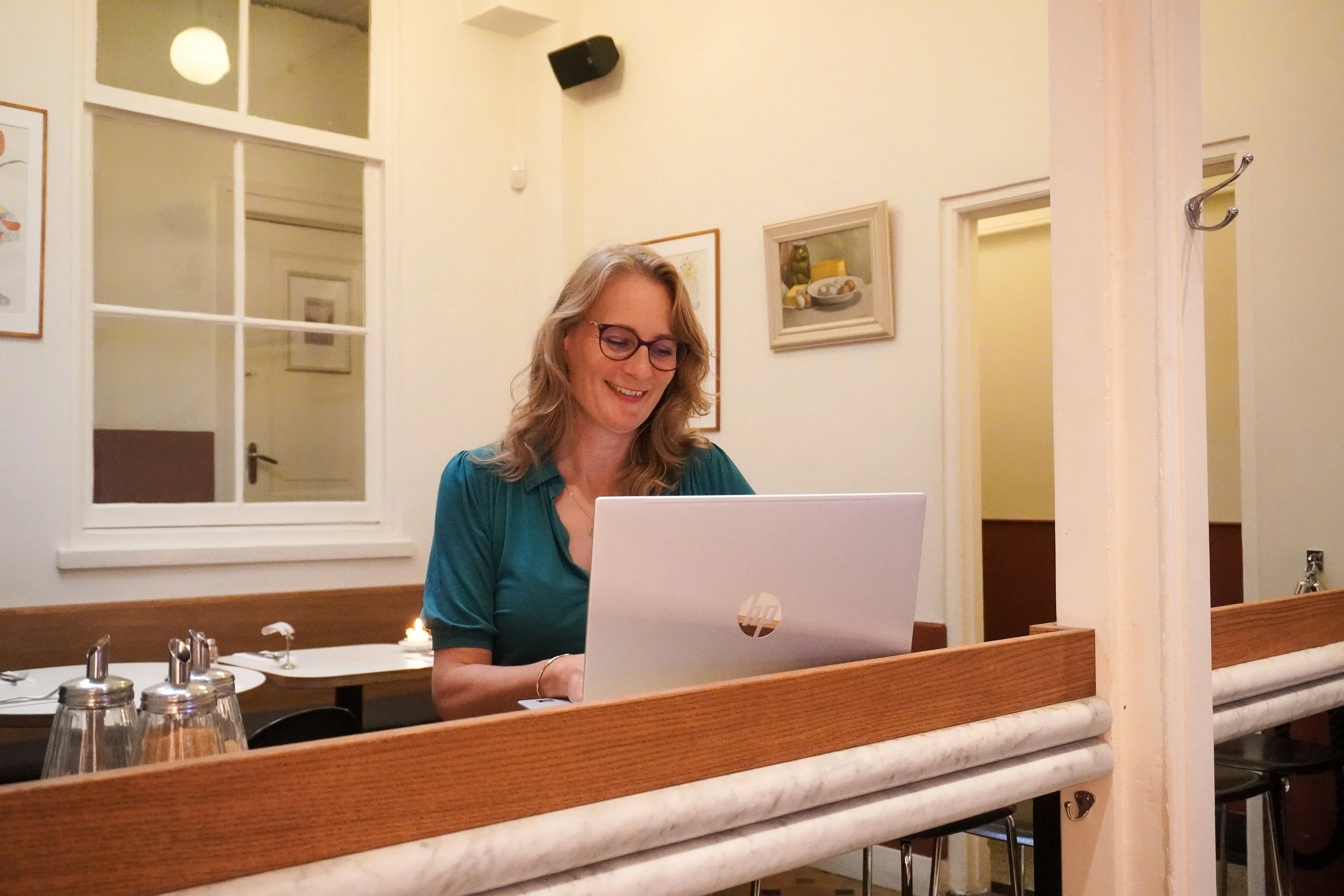 Een lachende vrouw met bruin haar en bril zit achter een witte laptop in een huiselijke omgeving, waarschijnlijk in een eetkamer of woonkamer, met kunst aan de muur en een raam op de achtergrond.