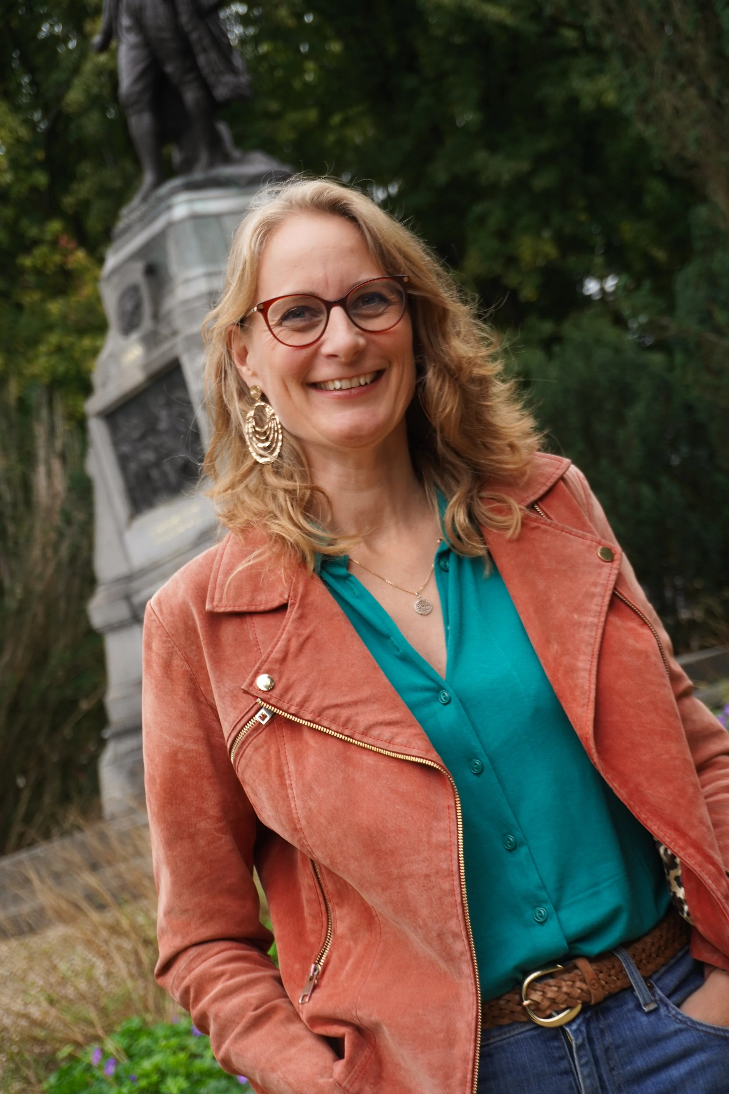 Vrouw met bruin haar en grote oorbellen, glimlachend, in een park met een monument op de achtergrond.