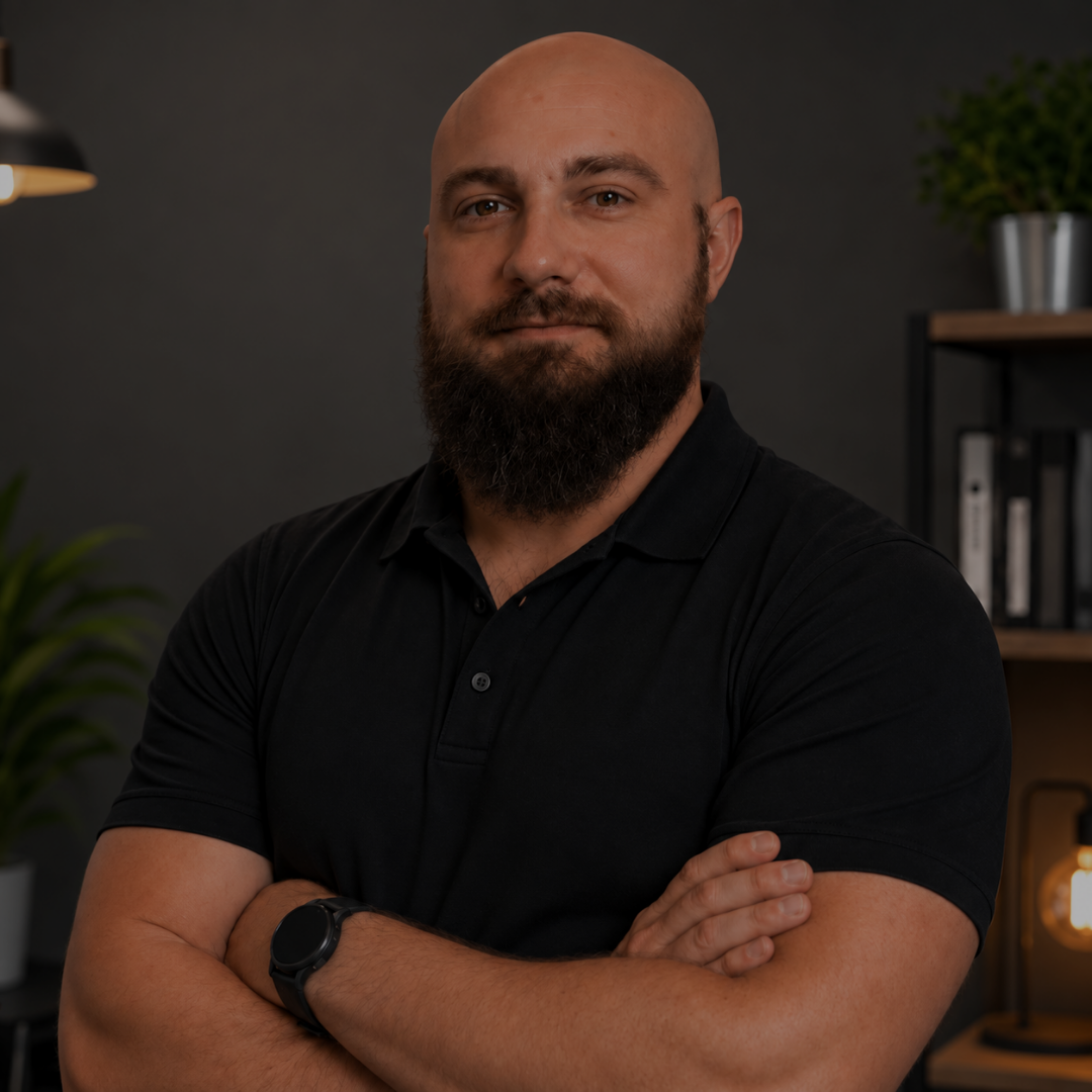 Homme avec une barbe portant un polo noir, bras croisés, dans un intérieur moderne.