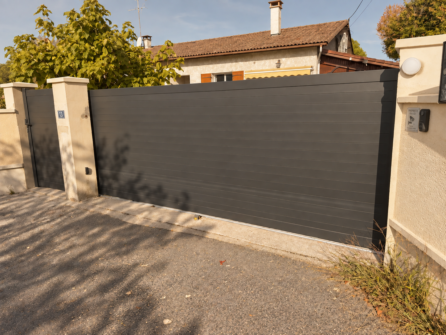 Une grande porte de clôture noire en métal avec un petit portail intégré, entourée de murs en pierre. Il y a également une maison en arrière-plan avec un toit en tuiles et des fenêtres avec volets en bois, ainsi que des arbres.
