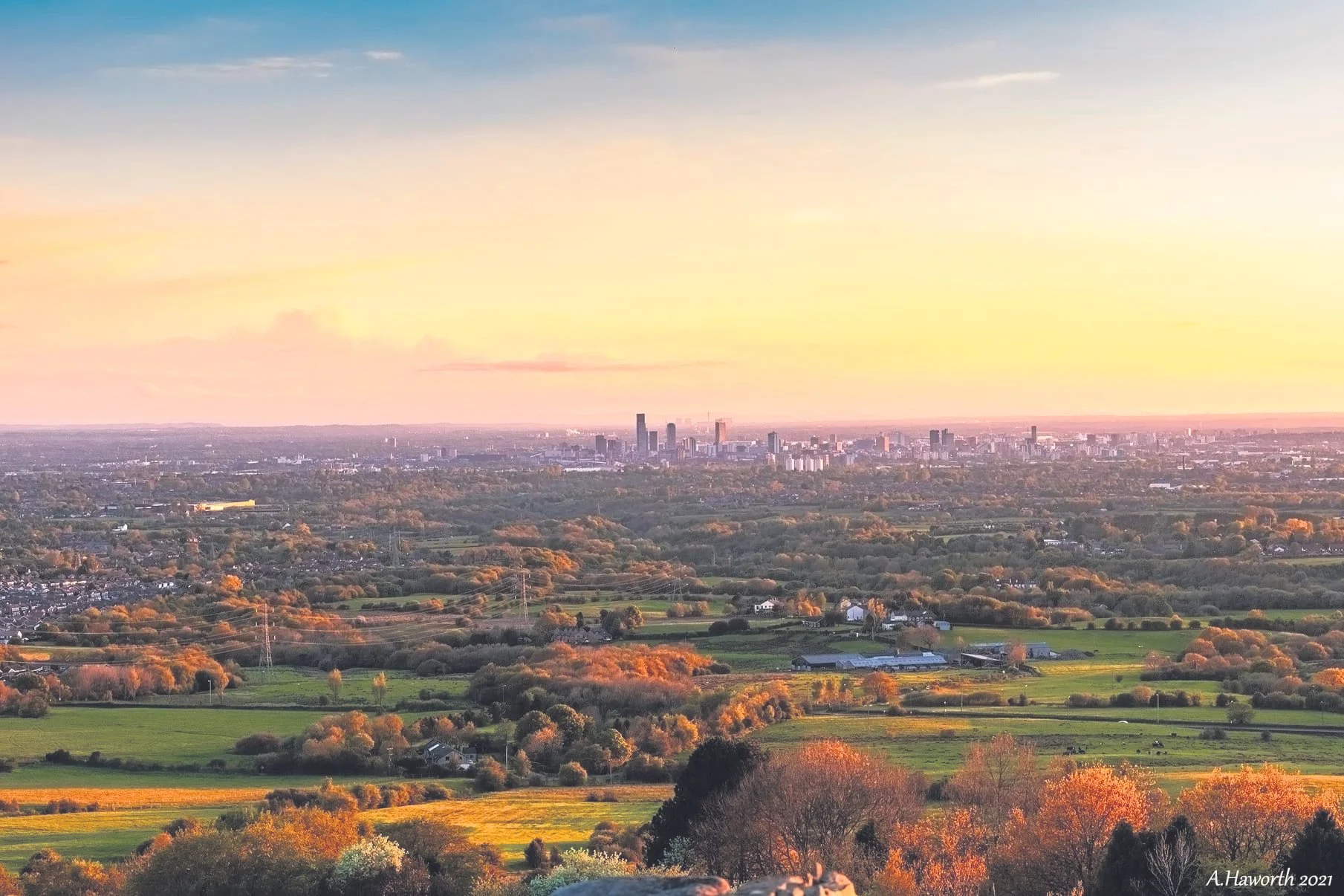 A panoramic view of a city skyline at sunset, with tall buildings in the distance and a foreground of green fields, trees, and small houses.