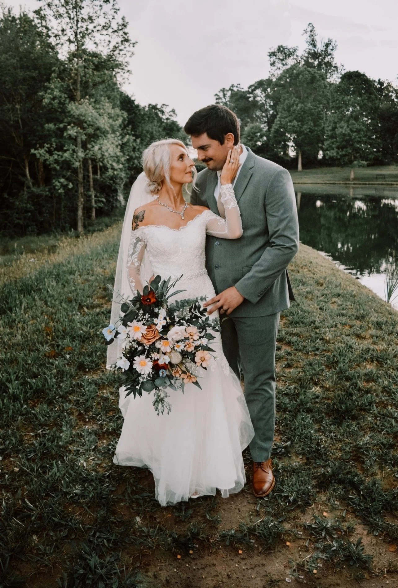 Bride and groom in wedding attire sharing an intimate moment by a pond, surrounded by trees, outdoor setting