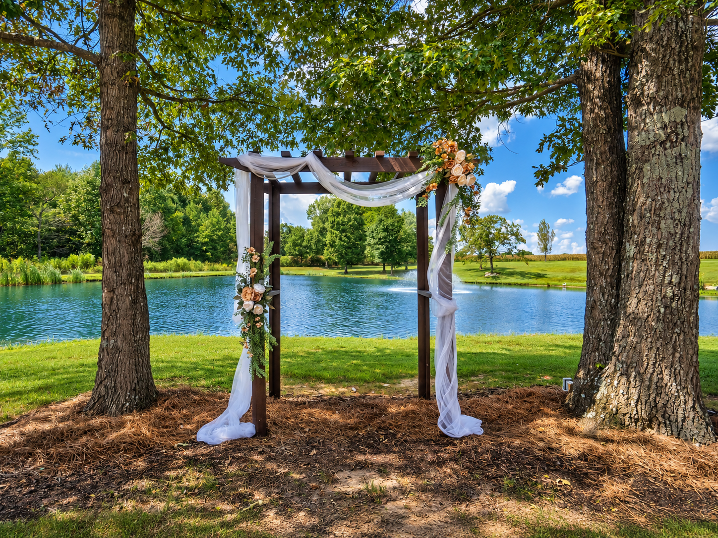 Wedding arch decorated with white fabric and flowers set in a natural outdoor setting near a lake, surrounded by trees under a blue sky.