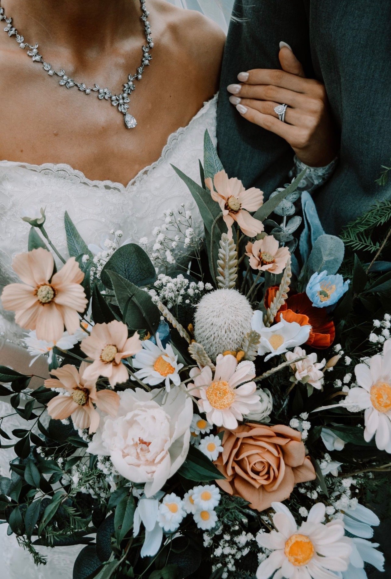 Close-up of a bride in a wedding dress holding a bouquet of flowers, wearing a diamond necklace and ring, with a man dressed in a suit next to her.
