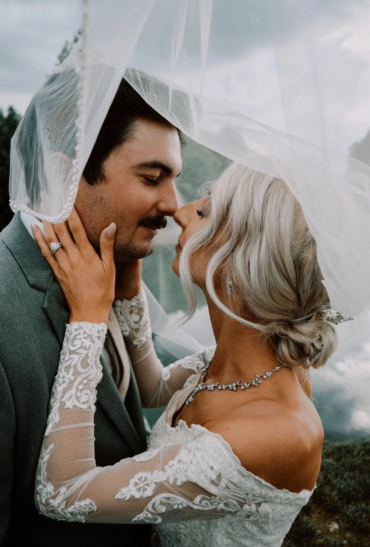 A bride and groom close-up, smiling with foreheads touching, under a sheer veil, outdoors with a cloudy sky and trees in the background.