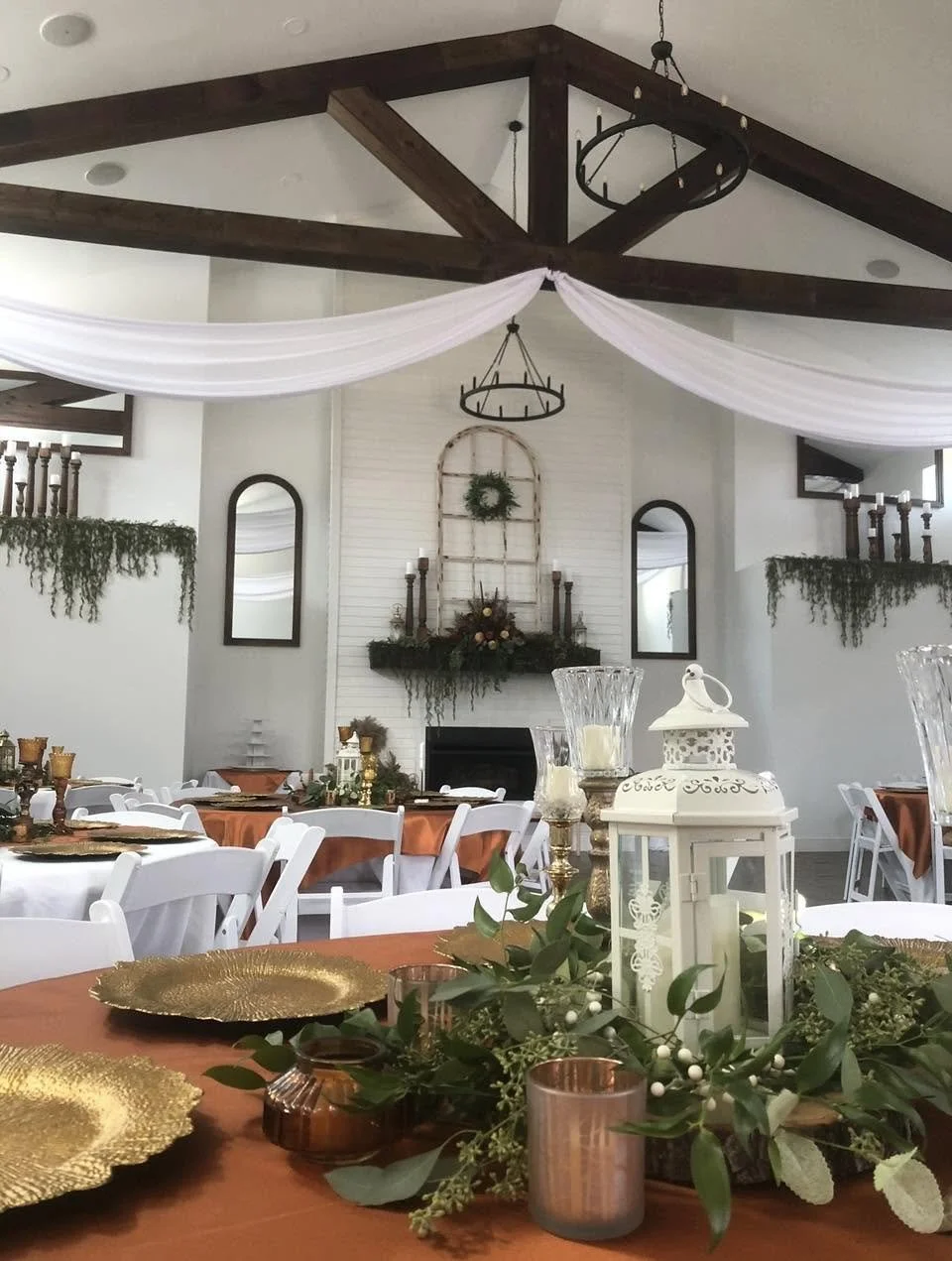 Indoor event space decorated for a celebration with tables covered in white and rust-colored tablecloths, gold chargers, greenery centerpieces, and candle holders, with draped white fabric and a central fireplace with holiday decorations.