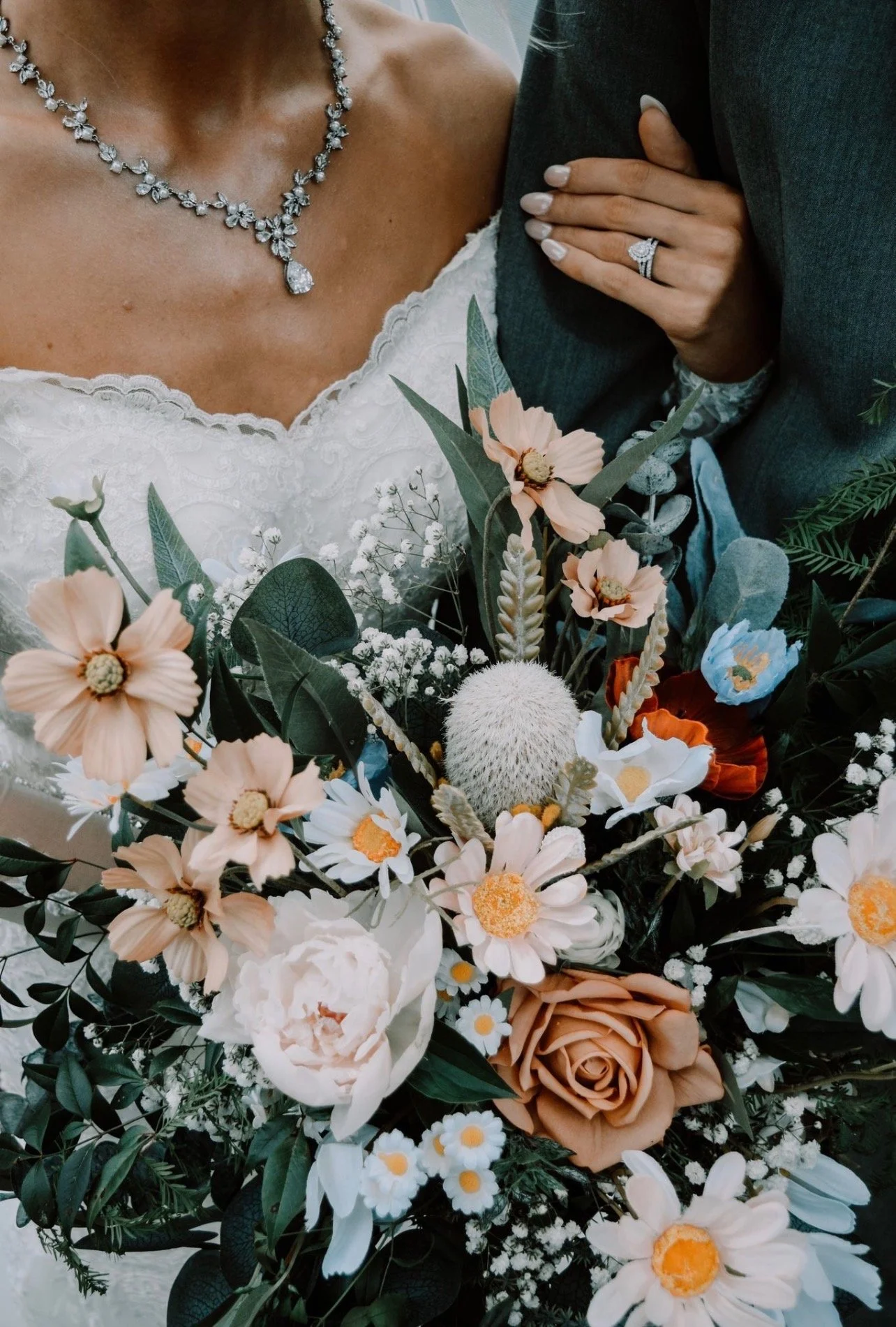 Close-up of a bride holding a bouquet of assorted flowers, including peonies, daisies, roses, and greenery, with a woman wearing a diamond necklace and a wedding ring beside her.