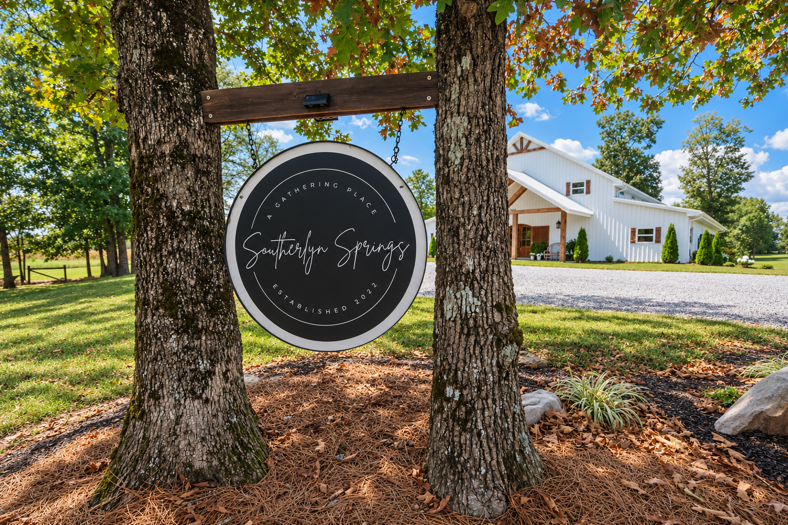 A round black sign hanging between two trees reading 'Southerly Springs, a gathering place, established 2022' in a rural setting with a white barn-style building and green trees in the background.