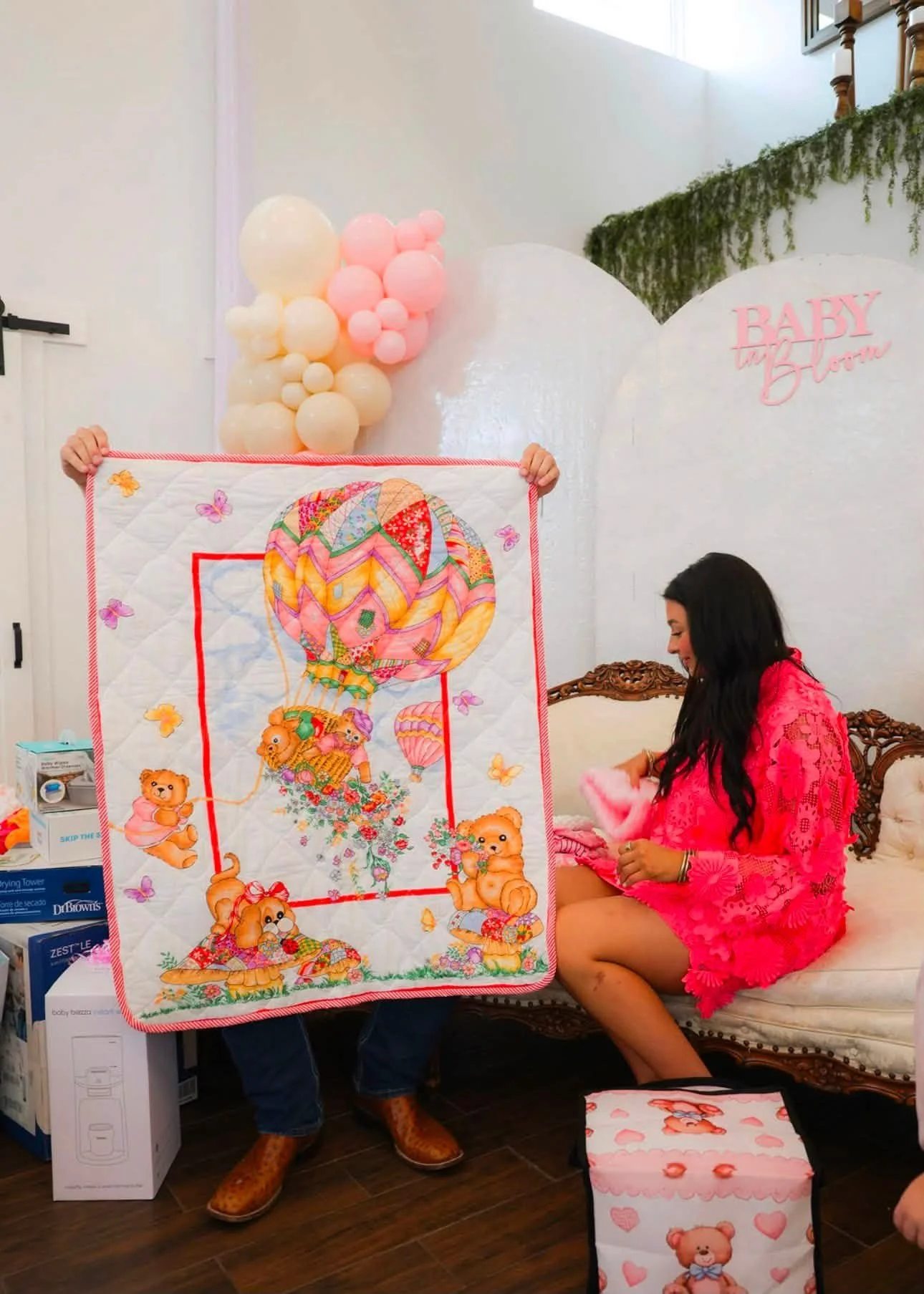 A woman in a pink dress sitting on a vintage sofa, looking at a pink blanket, with another person holding a colorful baby-themed quilt with a hot air balloon and teddy bears in front of her. Behind them, there are pink and cream balloons and a sign that reads 'Baby on Board'.
