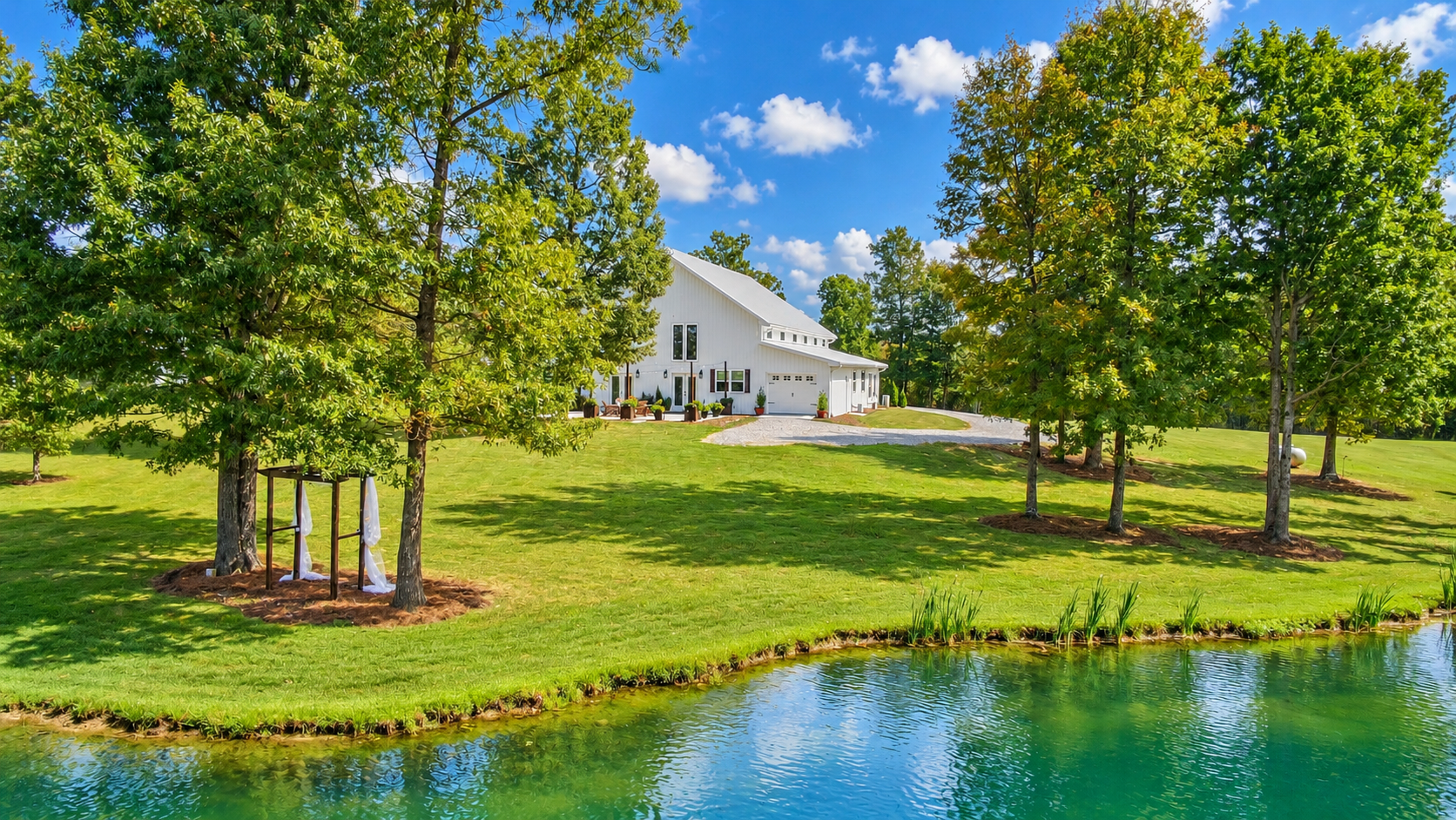 A white modern house with a side garage and large windows situated on a green lawn near a calm pond. Tall trees surround the house, some with draped white fabric, under a bright blue sky with scattered clouds.