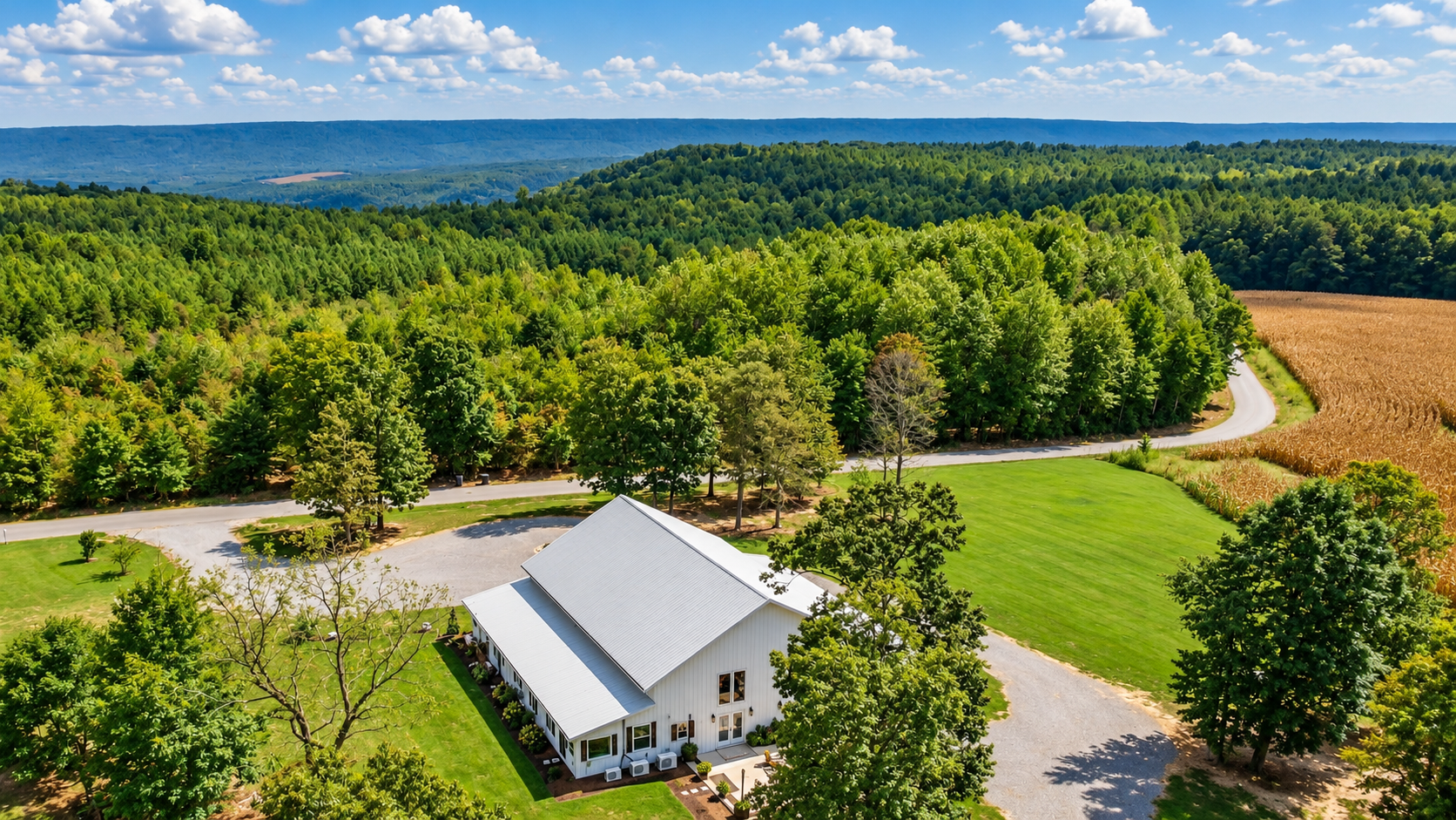 Aerial view of a white house with a gray roof surrounded by green trees, a well-maintained lawn, and fields of crops in a rural landscape, with hills in the distance under a partly cloudy sky.