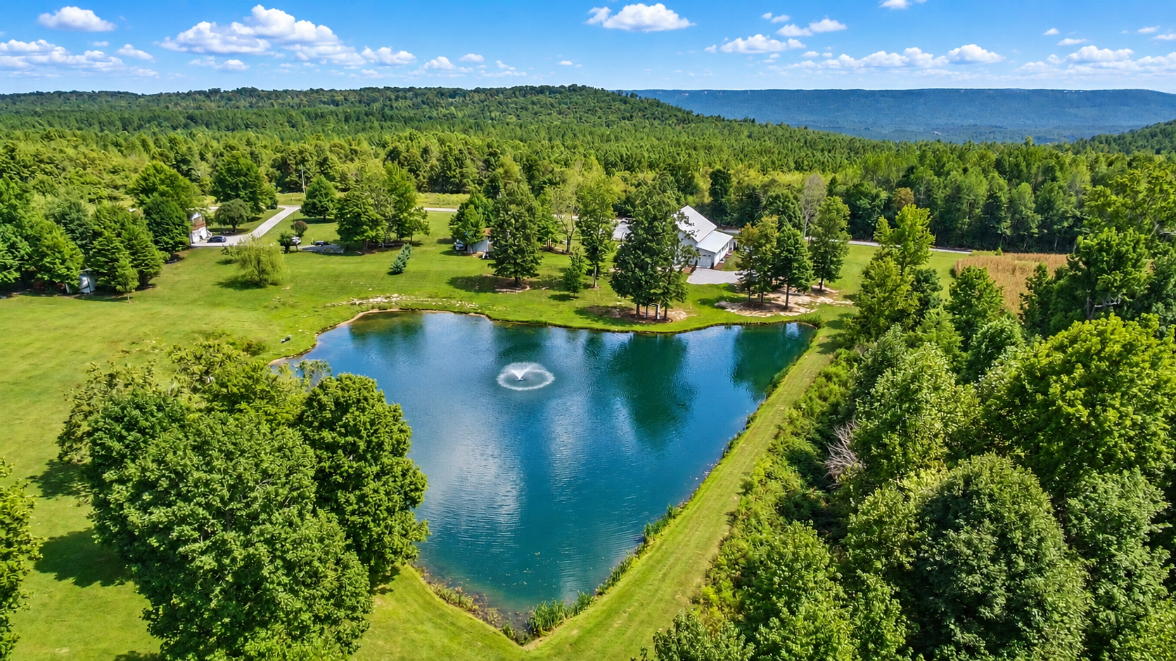 Aerial view of a small lake with a fountain in the center surrounded by green trees and open grassy areas, with houses and a wooded forest in the background under a partly cloudy sky.