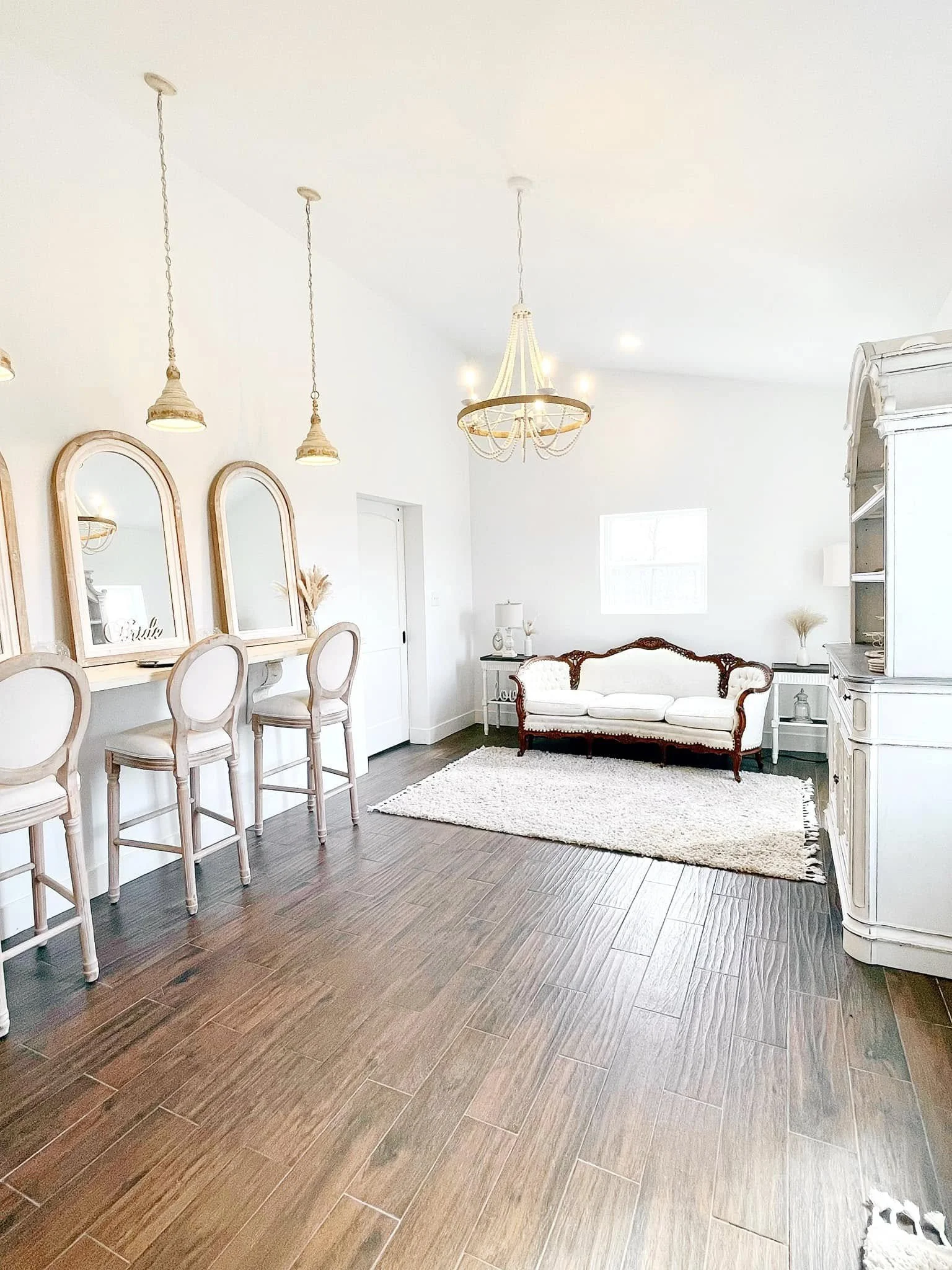 Living room with wooden flooring, white walls, and a white sofa with dark wood accents. There is a rug, a white side table with a lamp, and a vintage white cabinet. Three pendant lights and a chandelier hang from the ceiling. Four arched mirrors are 